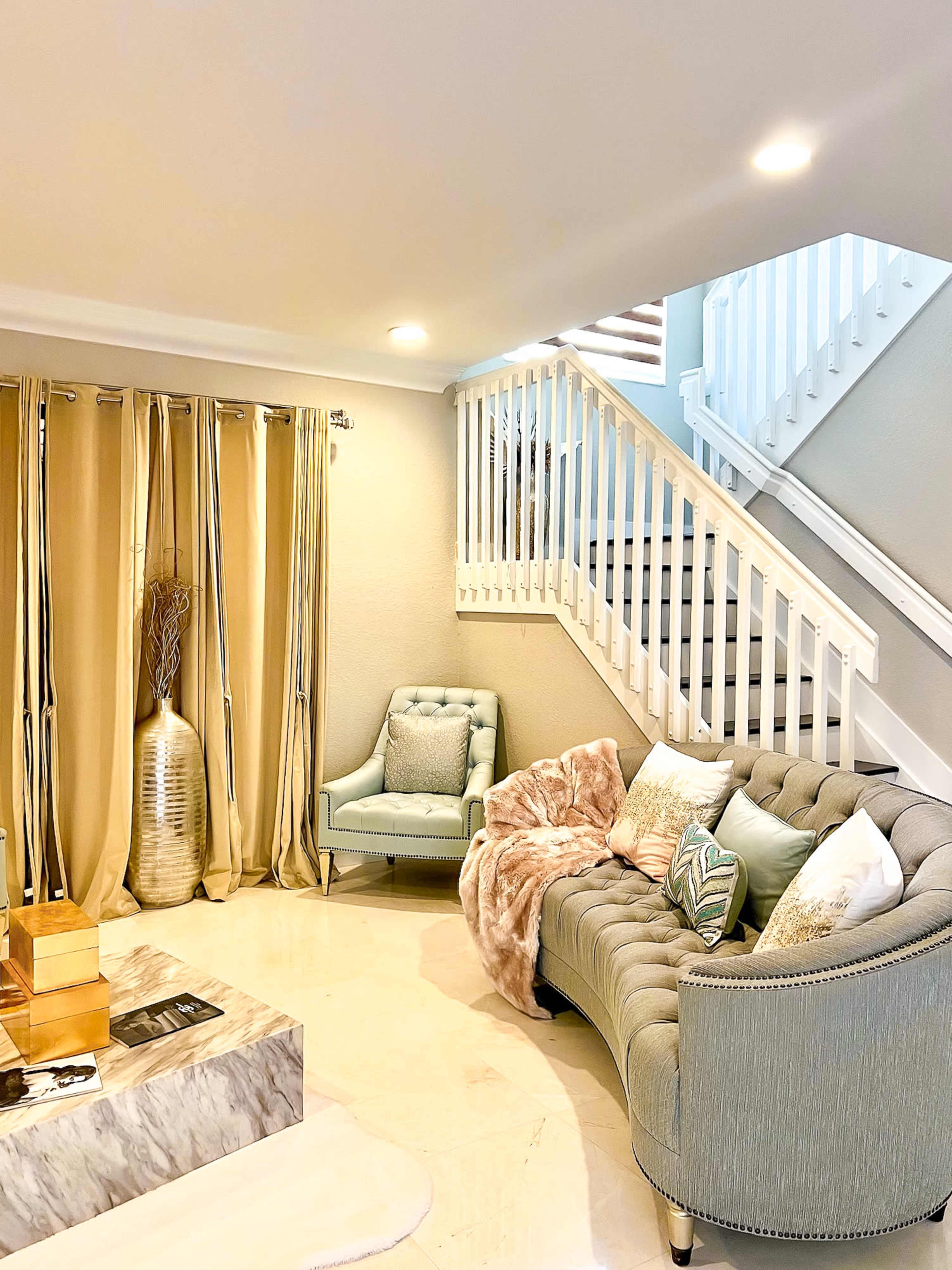 A living room features a gray tufted sofa, a light blue chair, a marble coffee table, and golden curtains, with a staircase visible in the background.