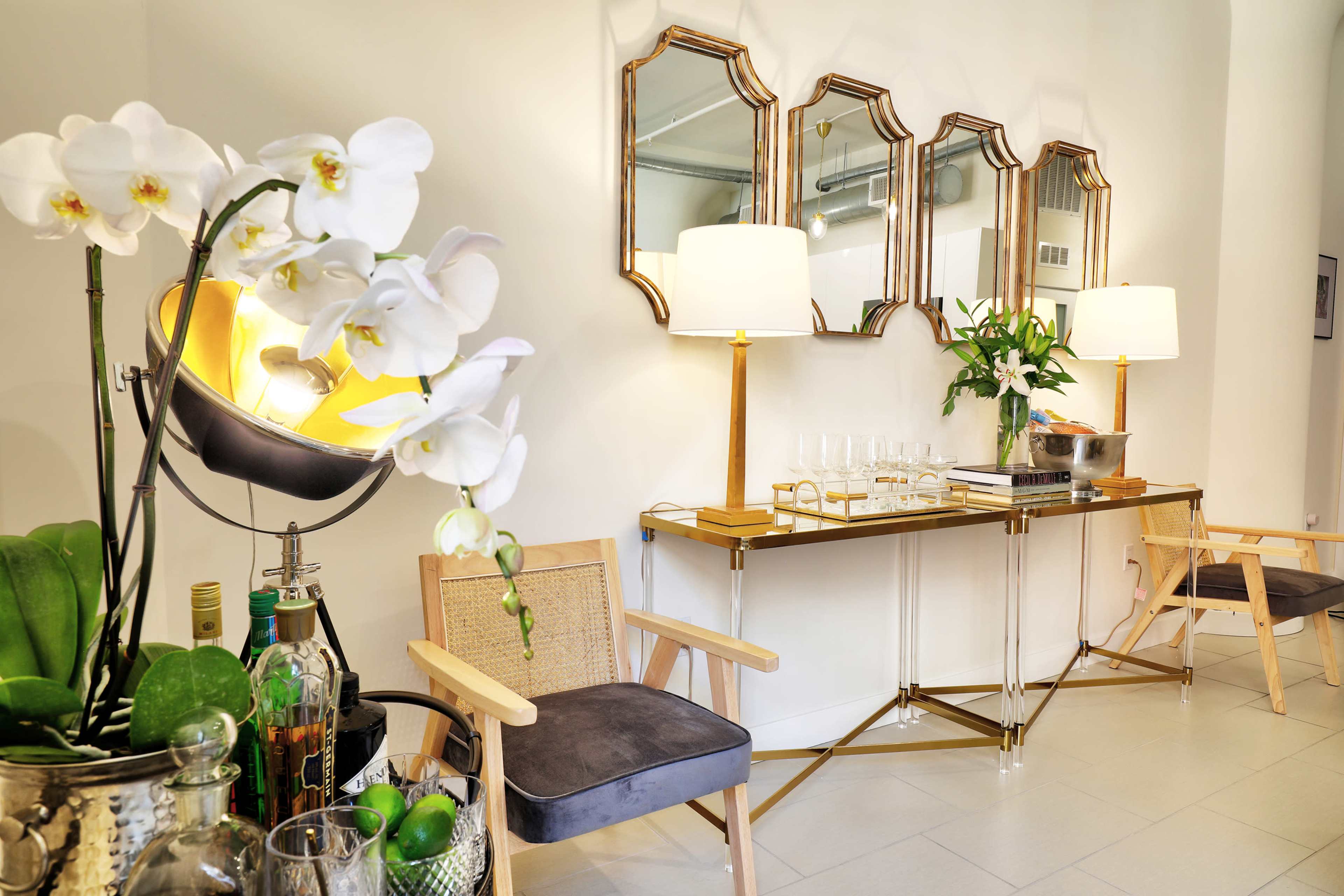 A well-decorated interior space features a sleek console table with three decorative mirrors above it, flanked by two wooden chairs and a decorative arrangement of orchids and bar accessories.