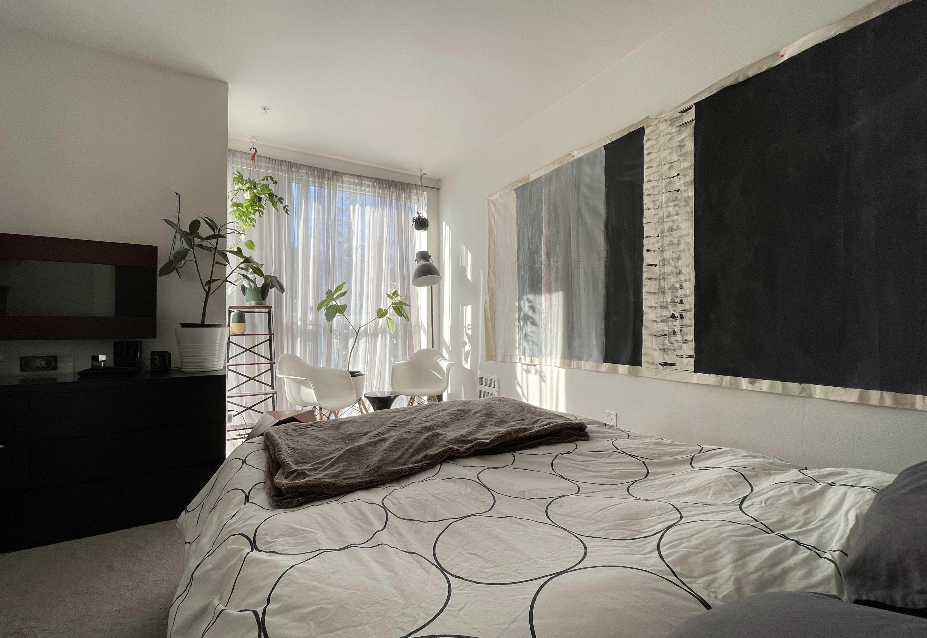 A bedroom features a bed with a patterned duvet, a television stand, and large windows with potted plants, leading to a small seating area with white chairs.