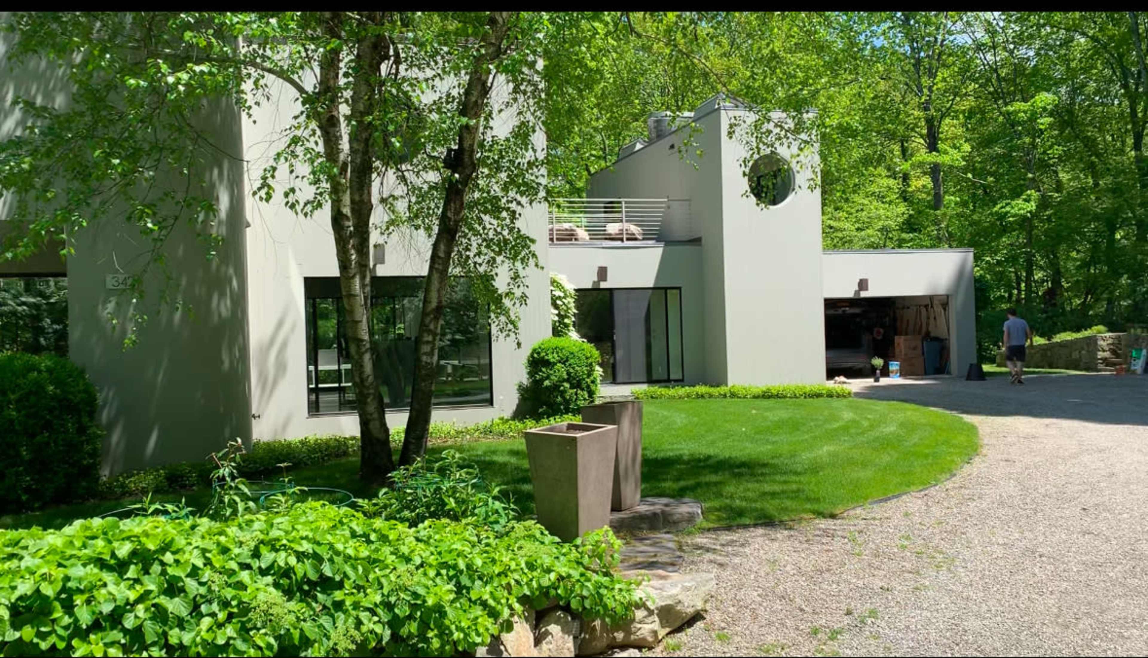 A modern, two-story house with large windows and a circular feature is set among green trees and a landscaped yard.