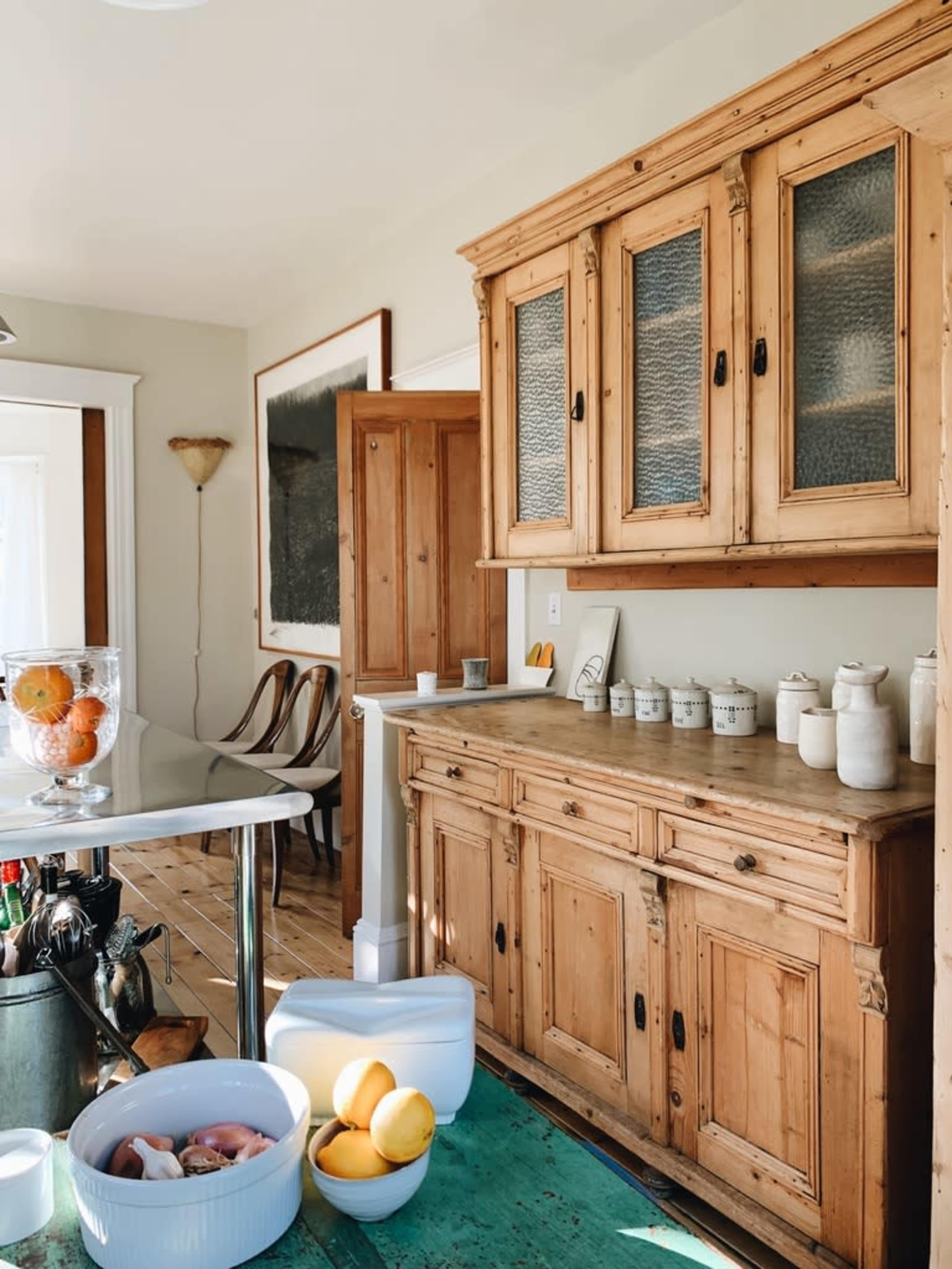 A rustic kitchen features a wooden sideboard with glass-paneled doors, a white table, and bowls of fruit on a green floor.