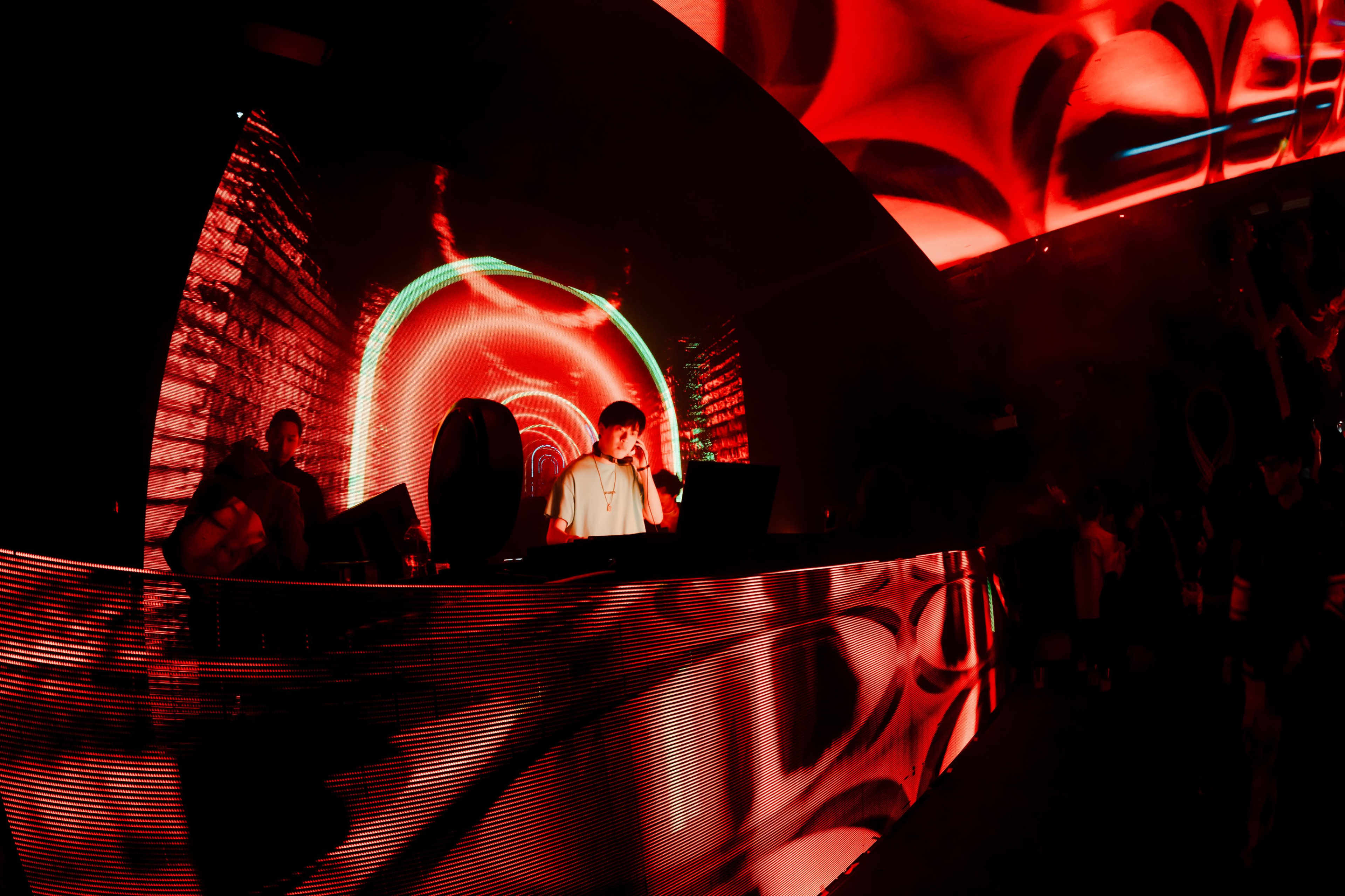 A DJ performs behind a large illuminated console with vibrant red and green lighting in a crowded nightclub.