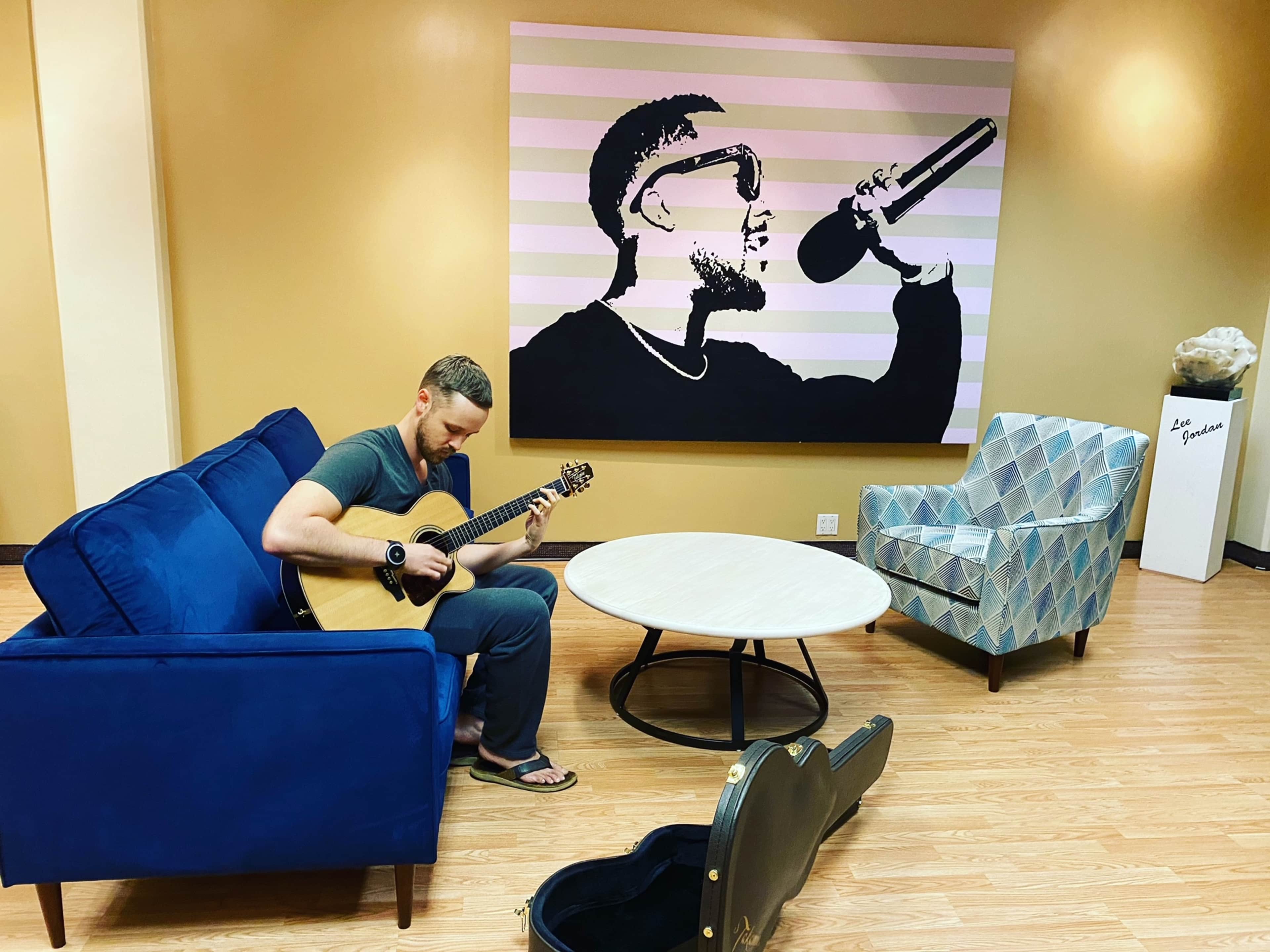 A man sits on a blue couch playing an acoustic guitar in front of a mural of a singer on the wall.