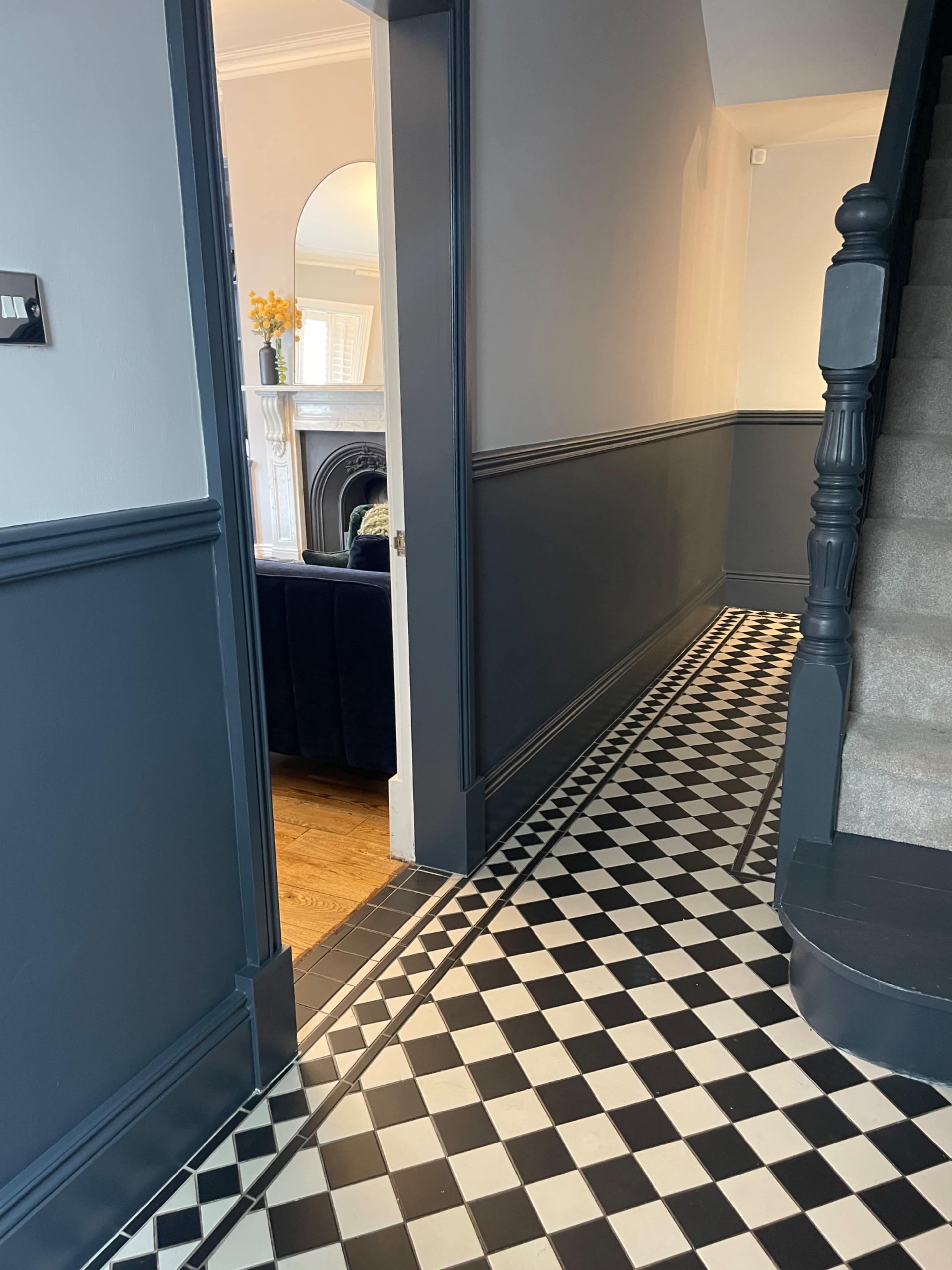 The image shows a hallway with a patterned black and white tiled floor, leading to a staircase and a living room visible through an open door.