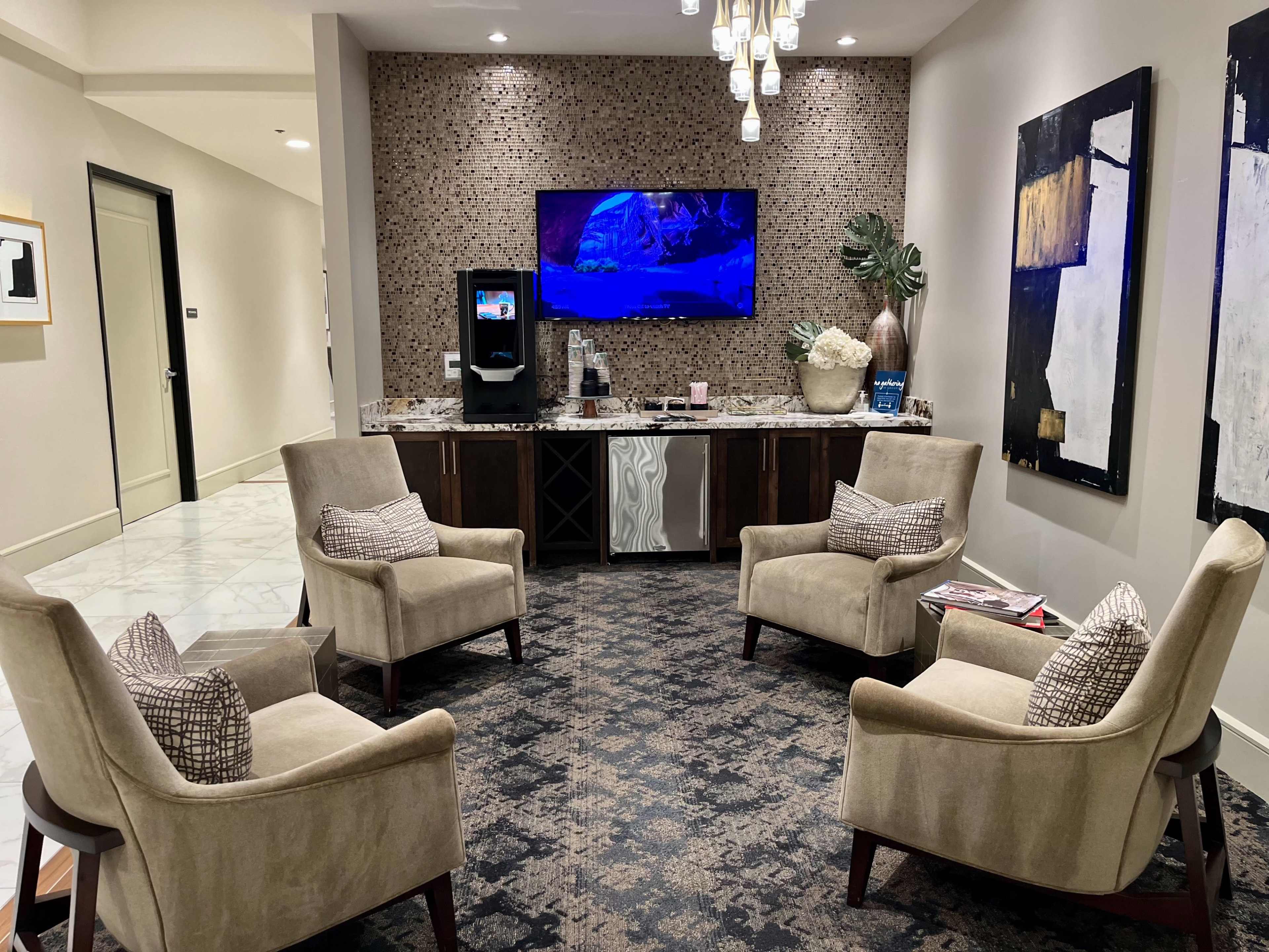 A waiting area with four beige armchairs arranged around a central table, a wall-mounted television, and a refreshment station with decorative elements.