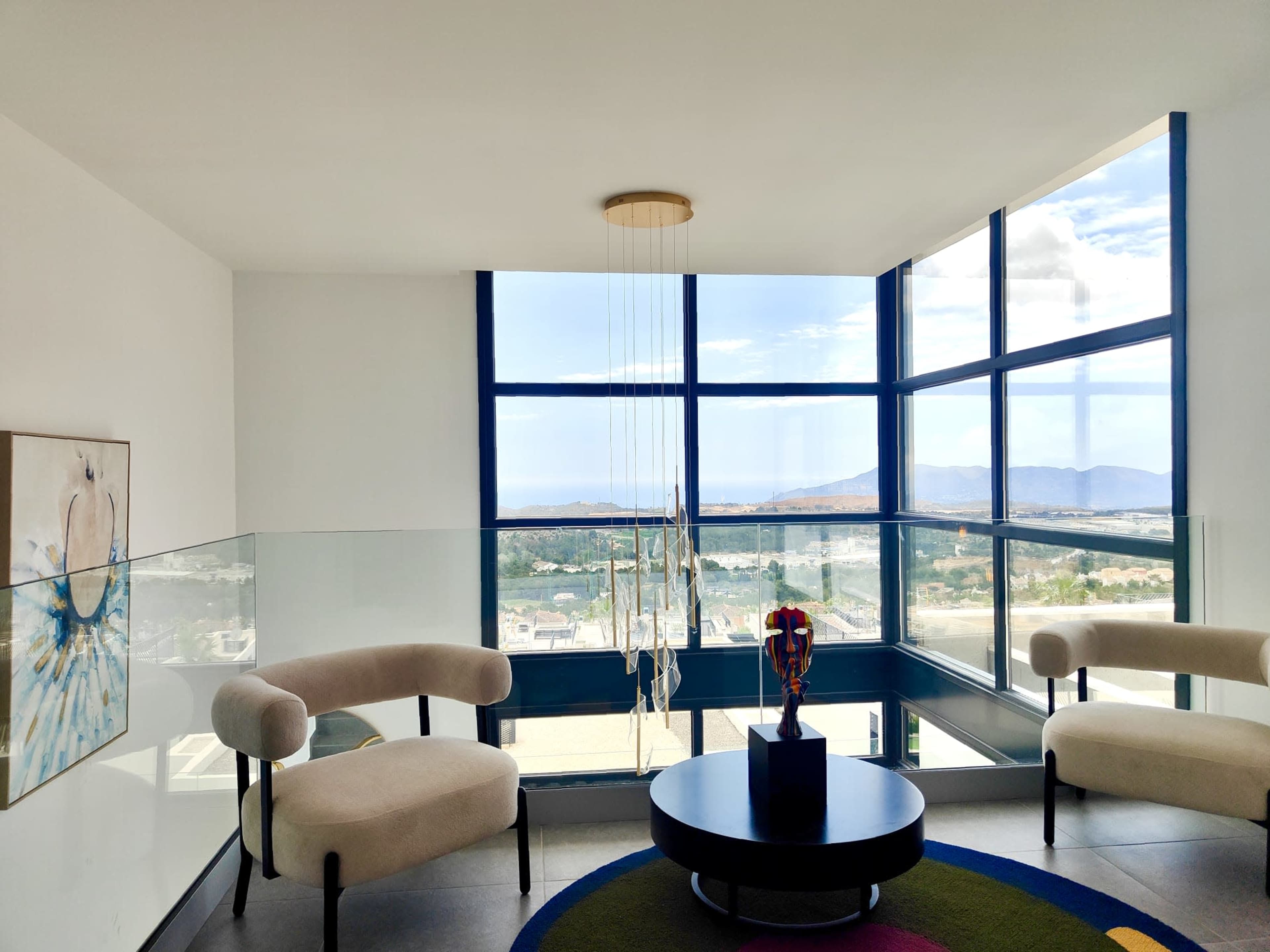 A modern living space features two upholstered chairs and a circular coffee table, with large windows displaying a view of hills and a cloudy sky.