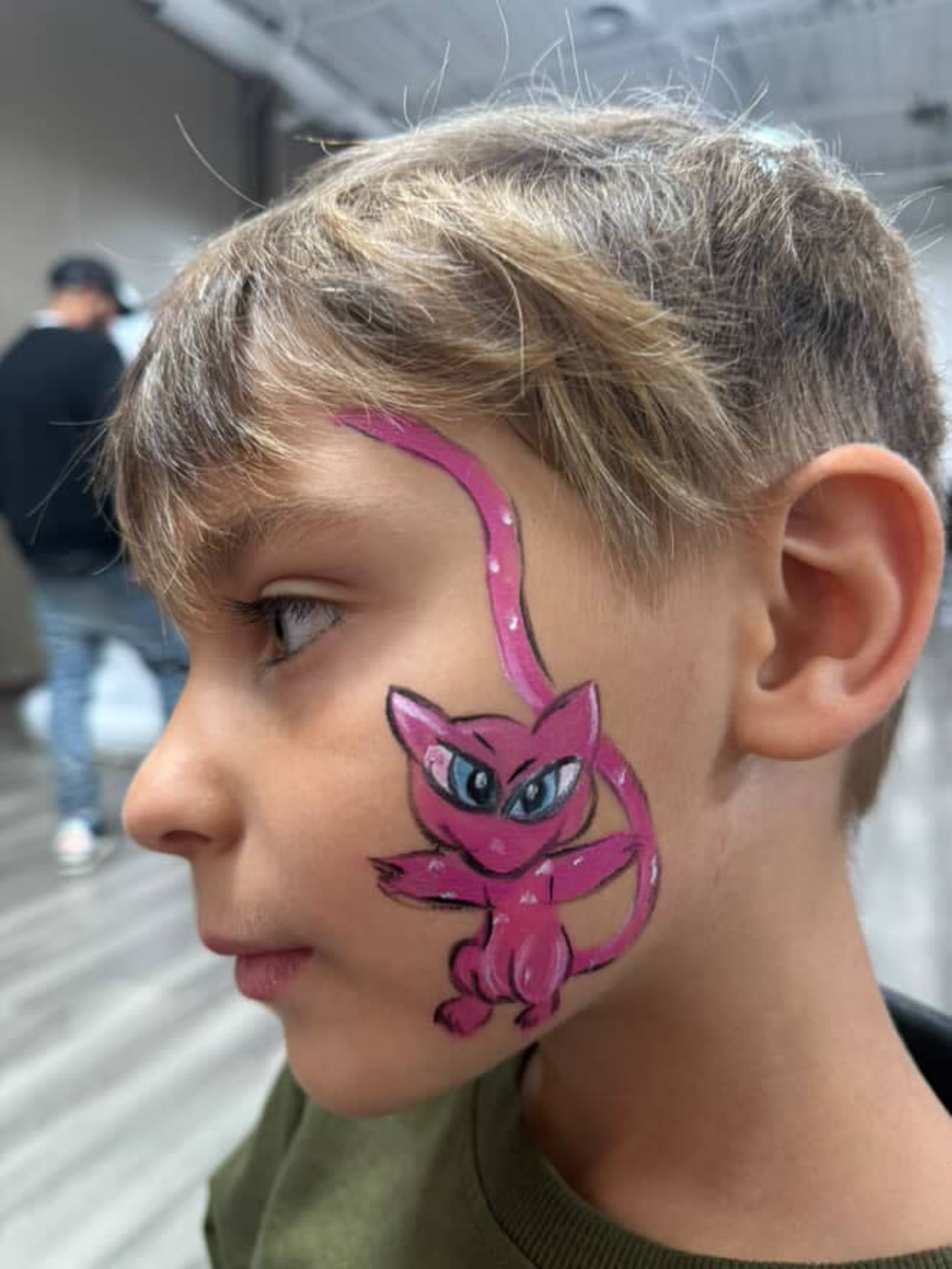 A young boy with a pink cartoon character painted on the side of his face stands in a brightly lit room.