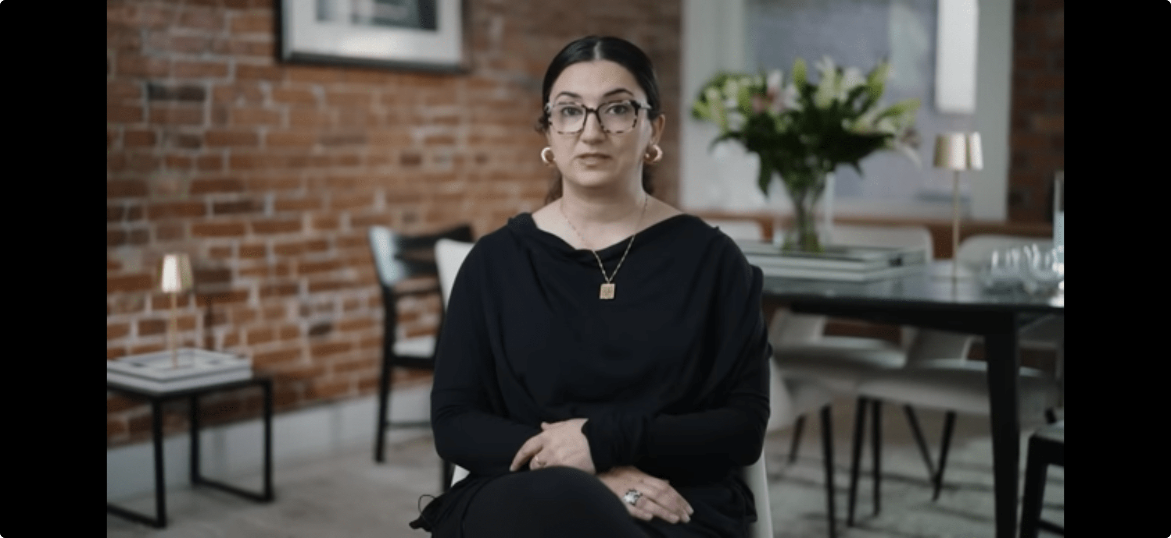 A woman with glasses is seated in a modern interior space featuring exposed brick walls and a dining table adorned with flowers.