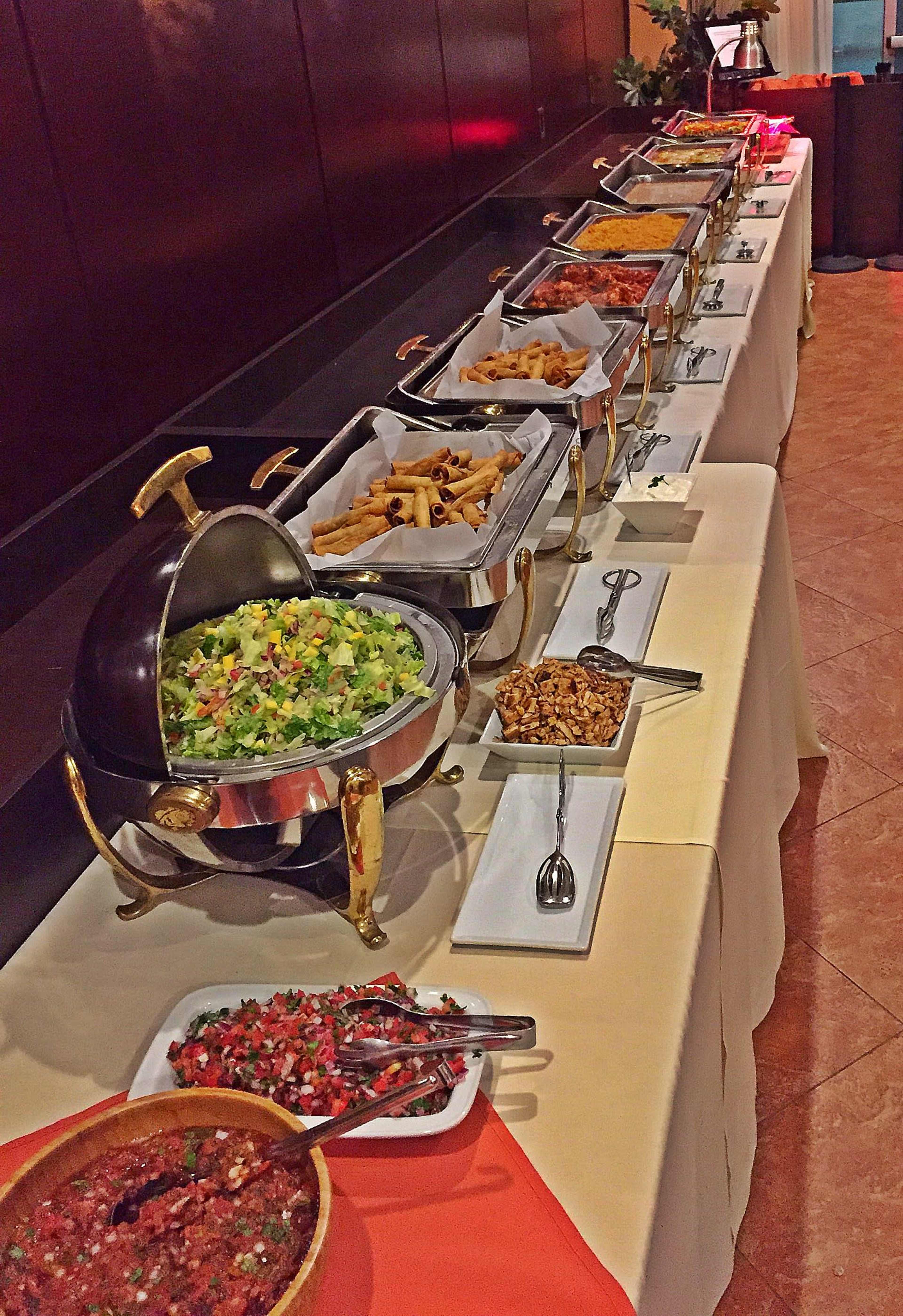 A long restaurant buffet table is set up with various dishes including salads, pasta, and toppings.