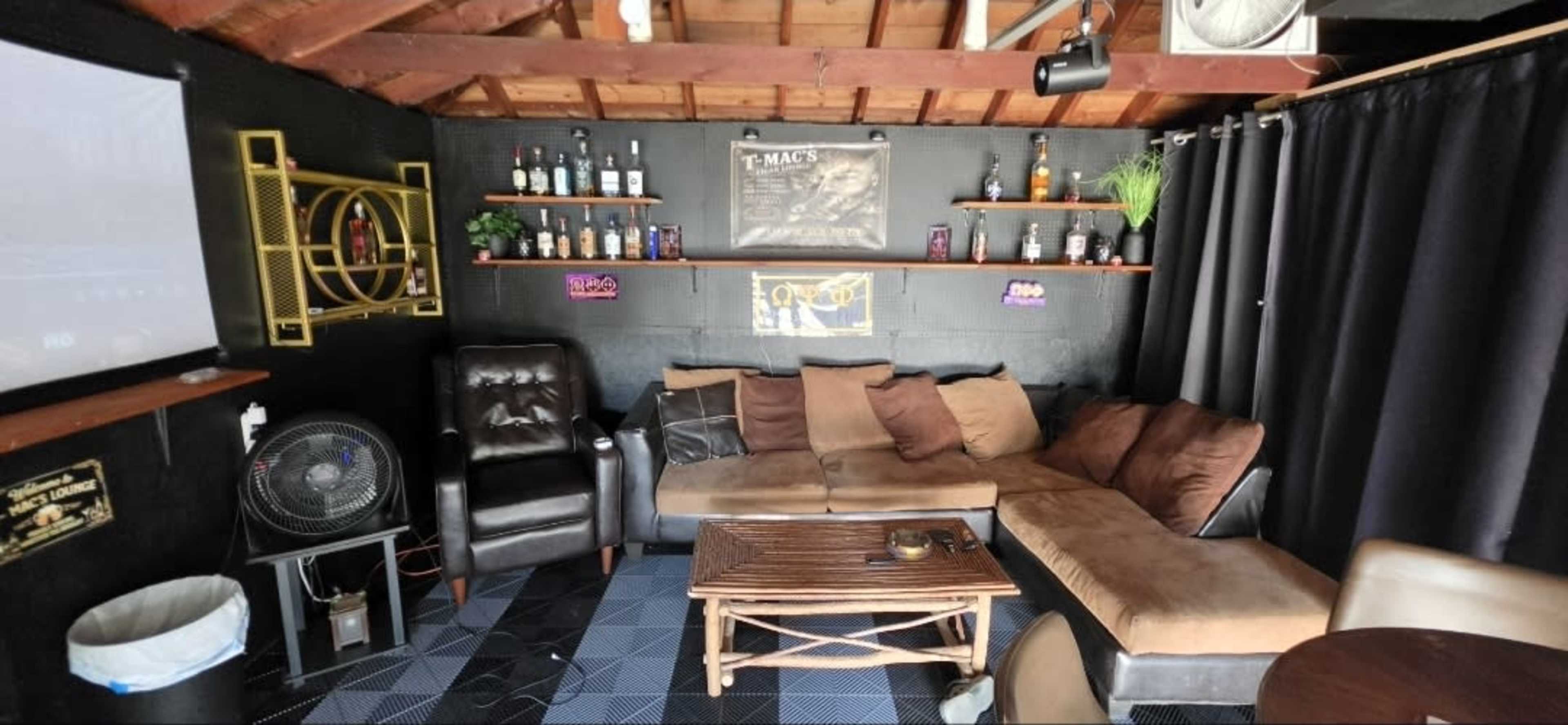 The image shows a cozy lounge area with a brown sectional couch, a recliner, a coffee table, and shelves adorned with bottles, all set against a dark wall with a projector screen.