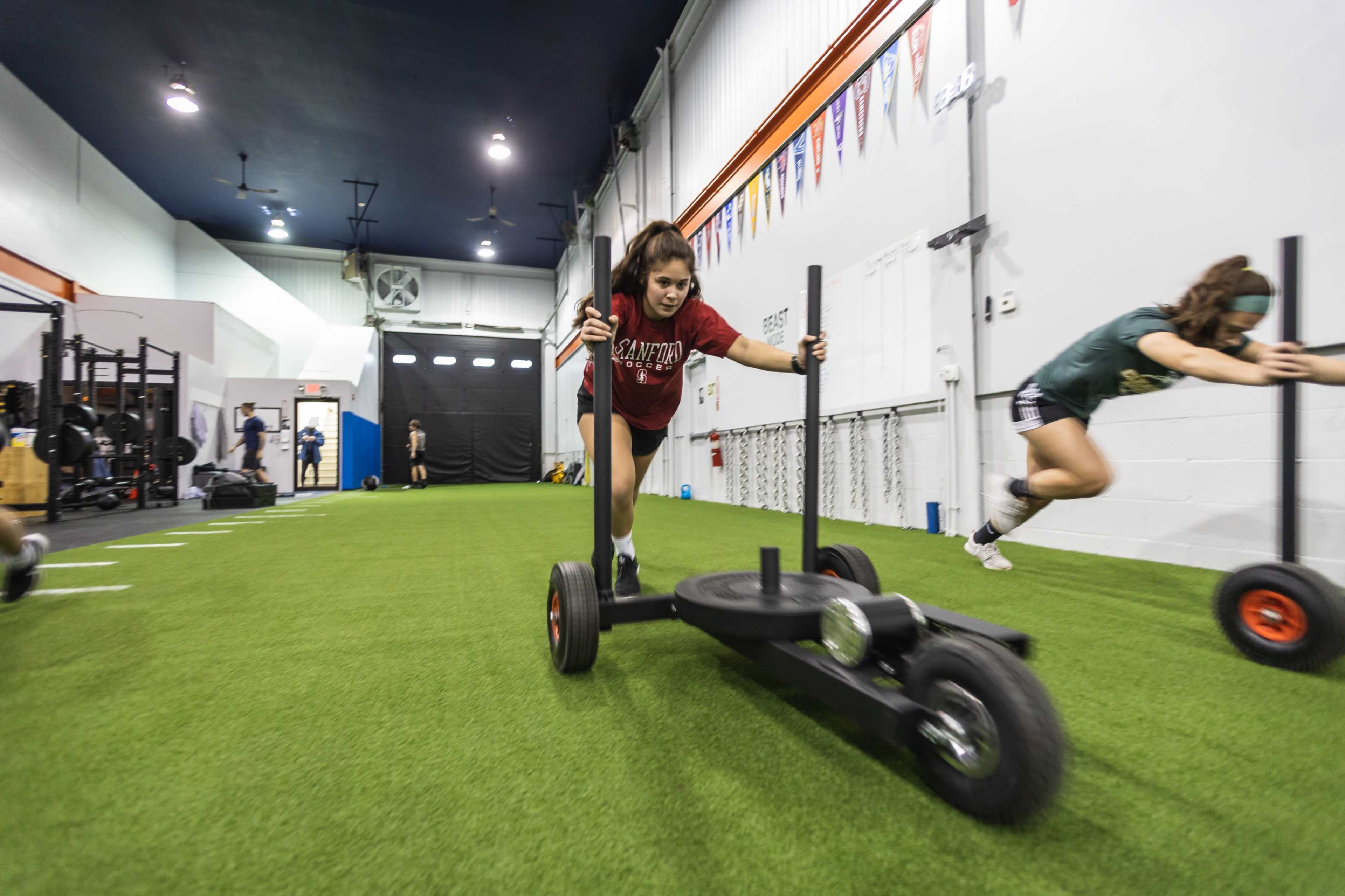 Two athletes are pushing sleds across a turf training area in a gym.