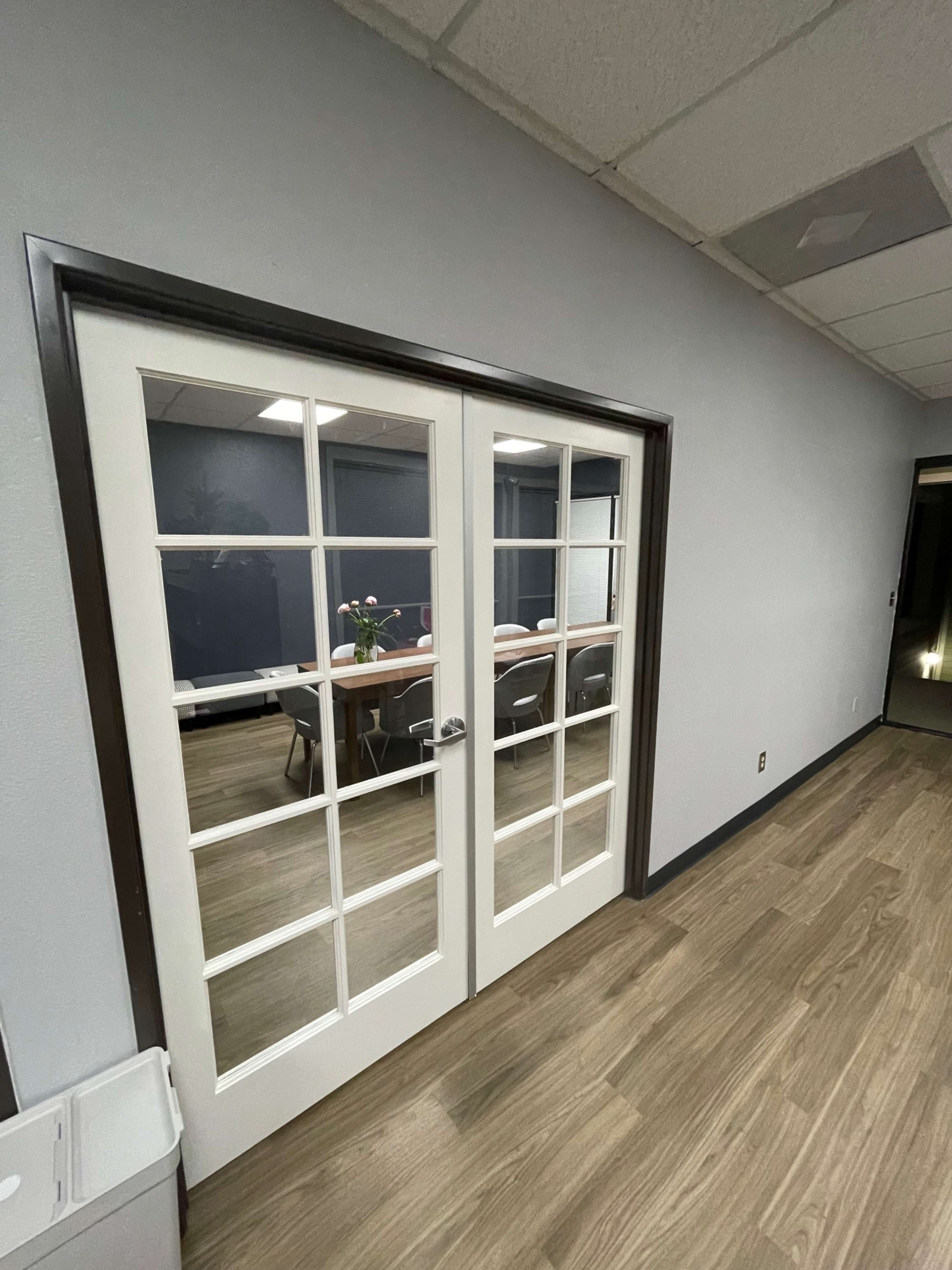 The image shows a set of glass-paneled double doors leading into a conference room with a long table and chairs visible inside.