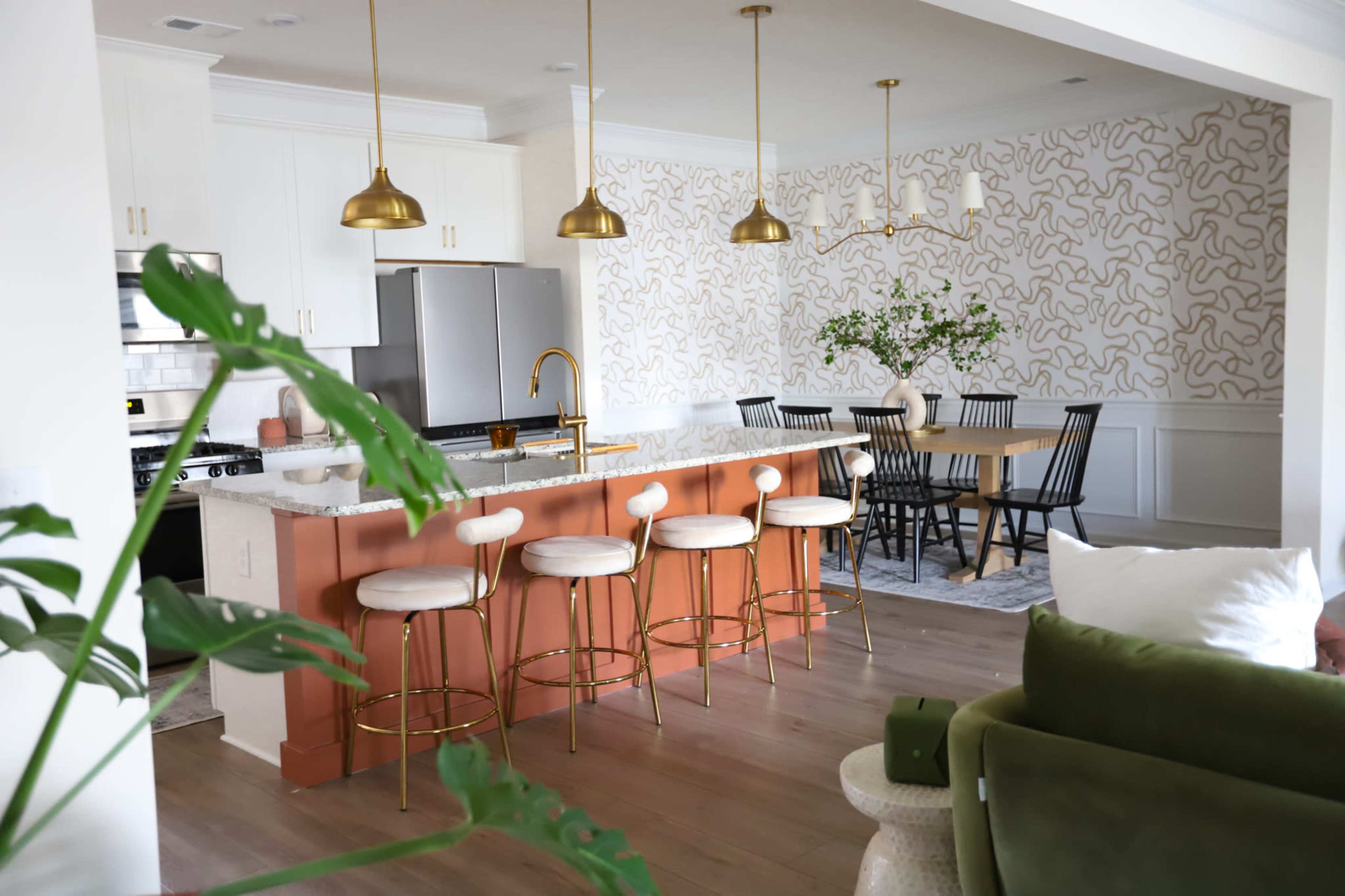 A modern kitchen and dining area featuring a light color palette, with a central island bar, gold fixtures, and a dining table surrounded by black chairs.