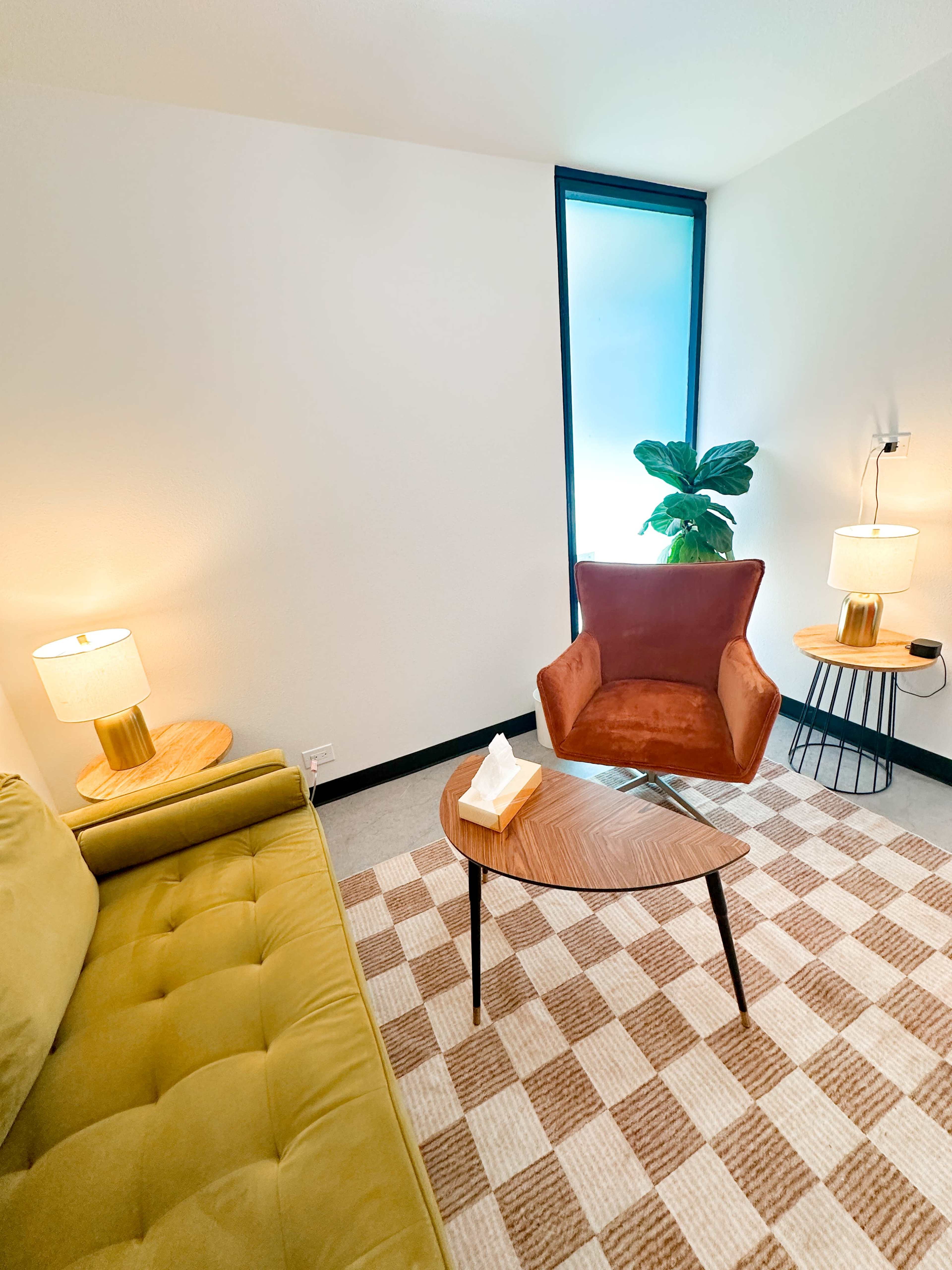 The image shows a cozy room with a green sofa, a round wooden coffee table, a patterned rug, and a potted plant near a window, illuminated by two lamps.
