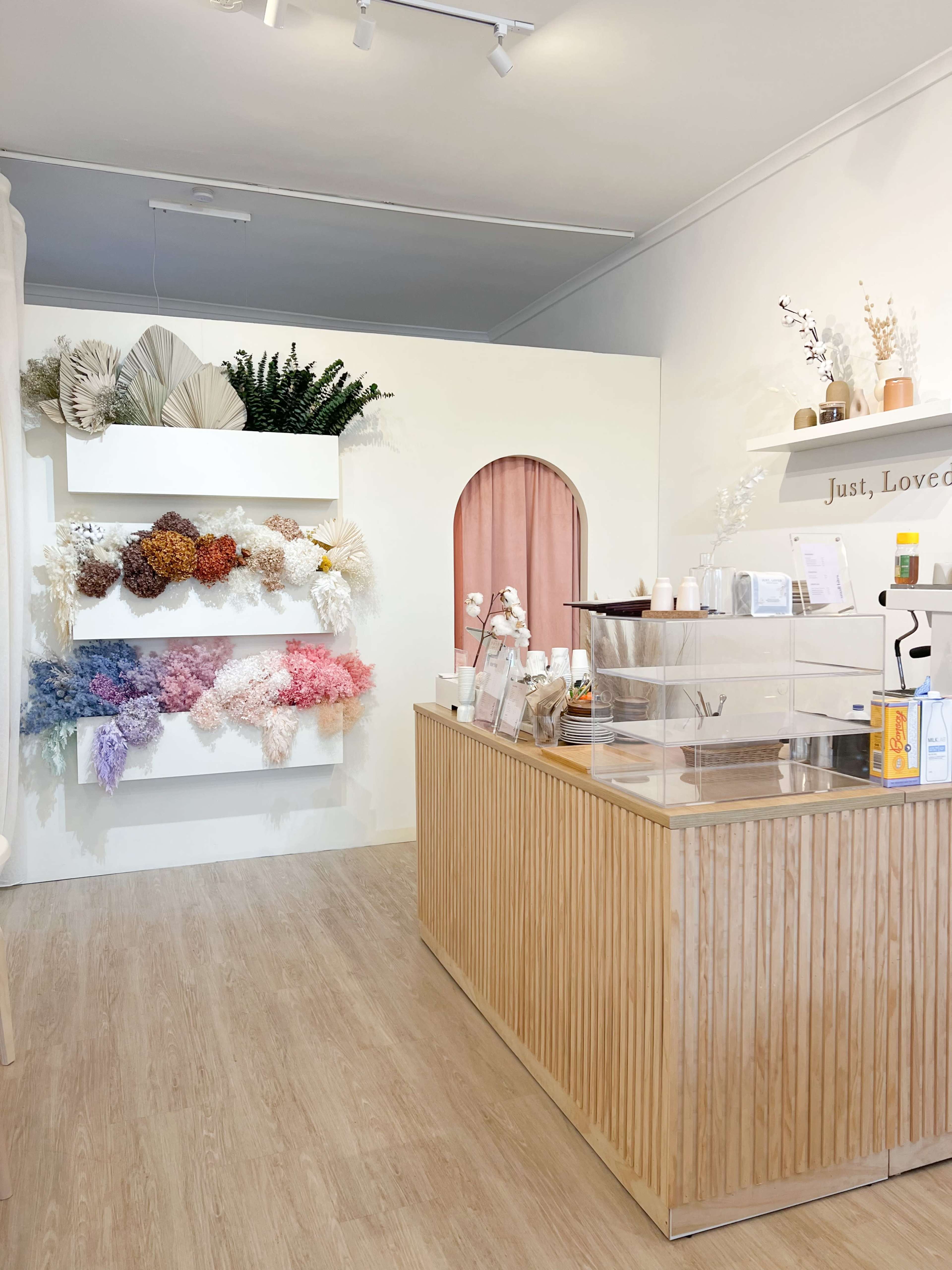 A shop interior with a wooden counter and a wall featuring arranged dried flowers in various colors, alongside a pink archway.