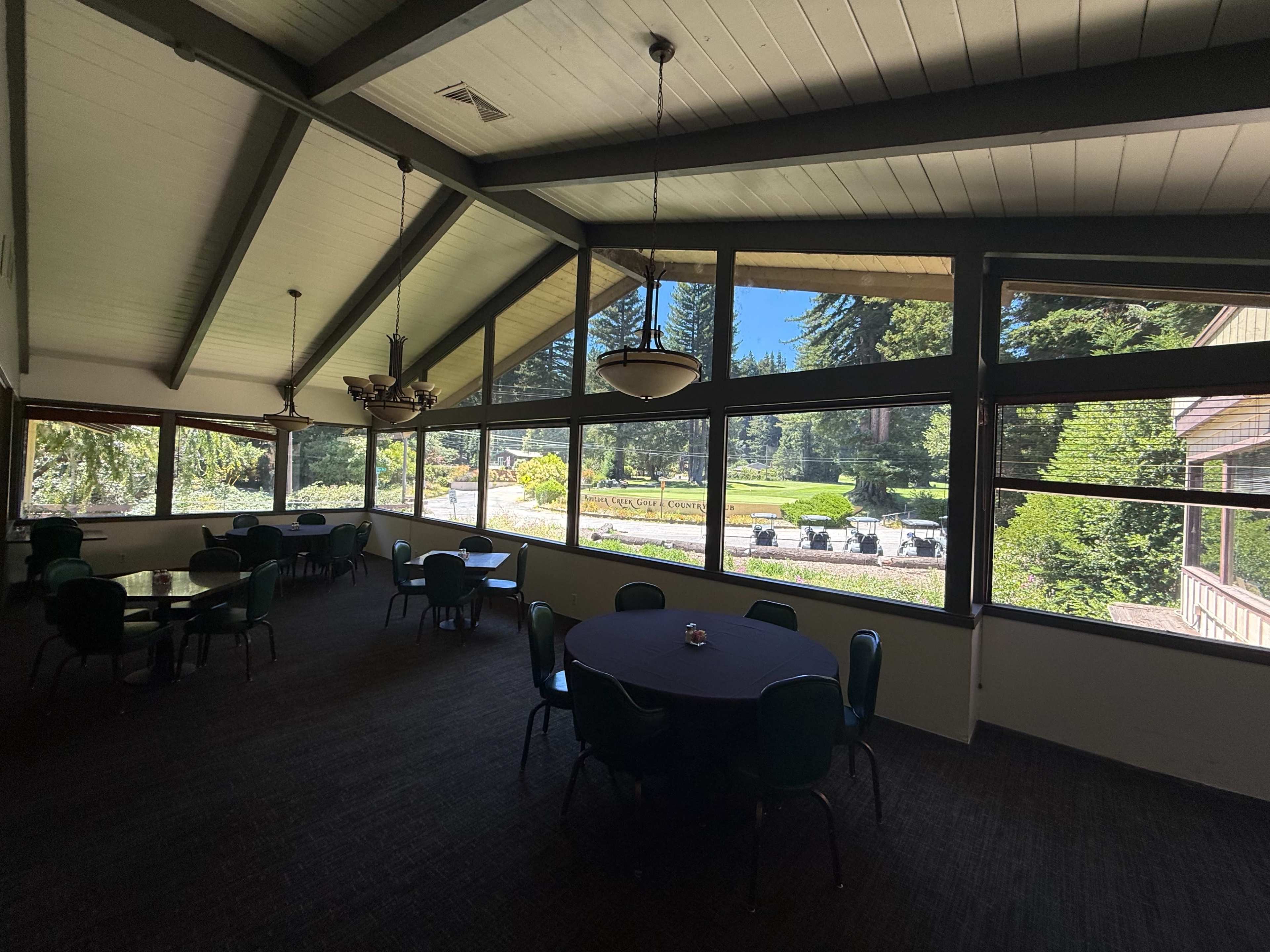 The image shows an interior room with high ceilings, featuring several round tables and chairs, and large windows overlooking a landscaped outdoor area.