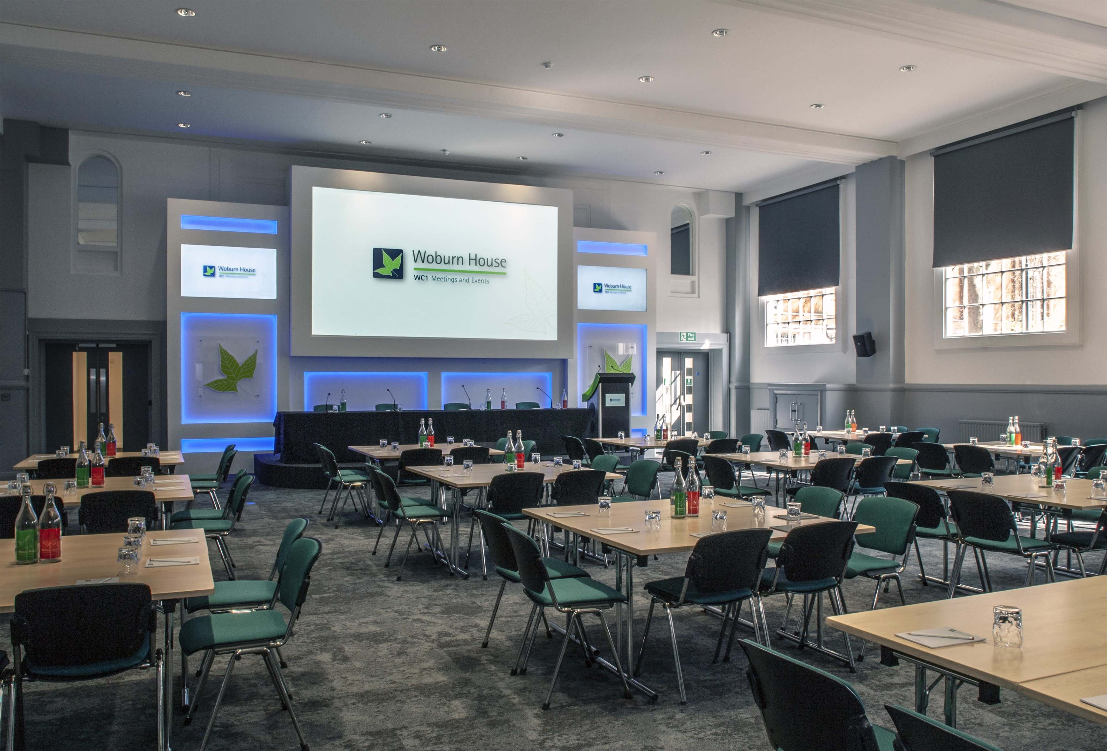 A conference room set up with multiple tables and chairs, featuring a large screen at the front displaying "Woburn House" with bottles of water and notepads on the tables.