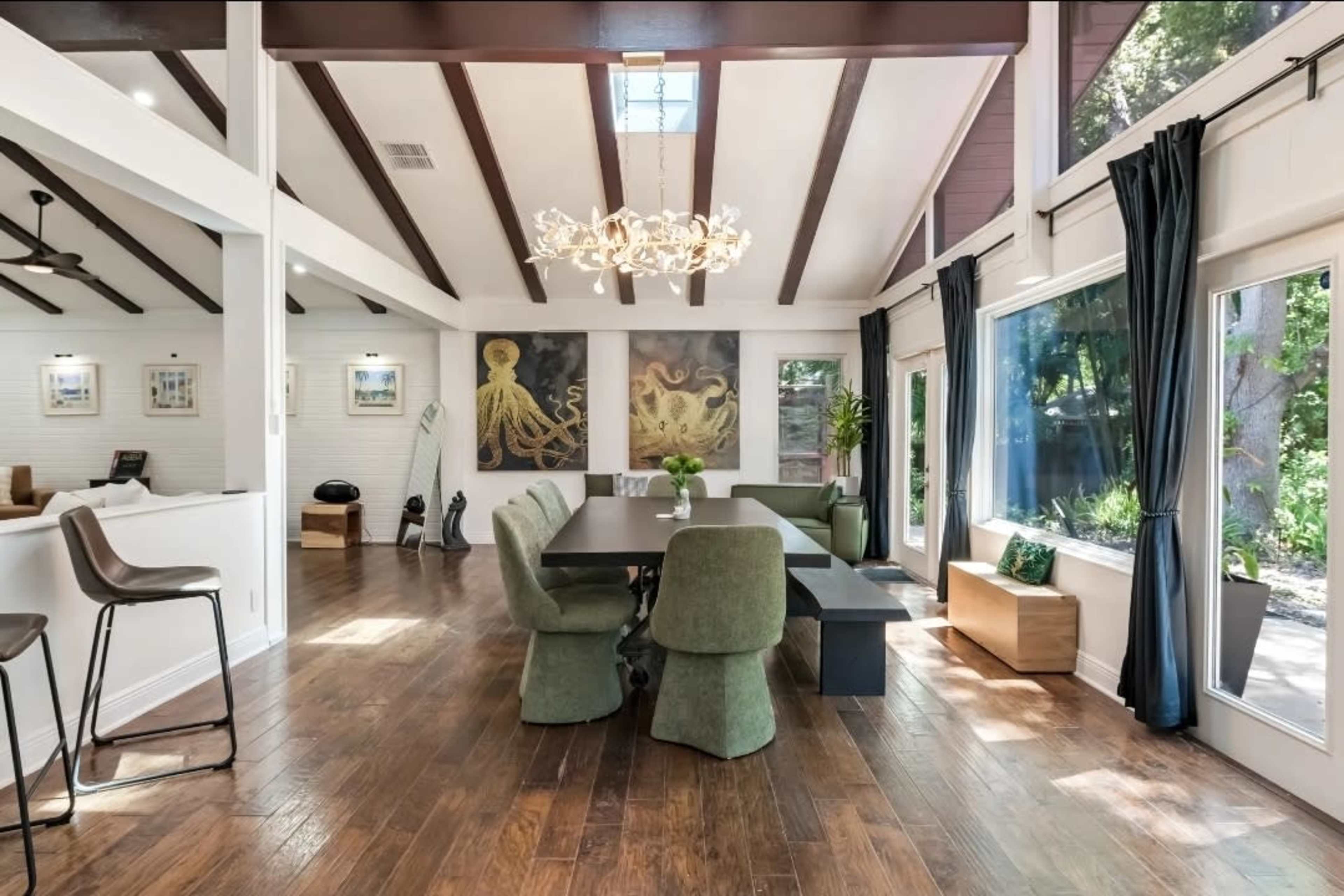 The image shows a modern dining area with a large table surrounded by green chairs, featuring a chandelier and artwork on the walls, with large windows allowing natural light.
