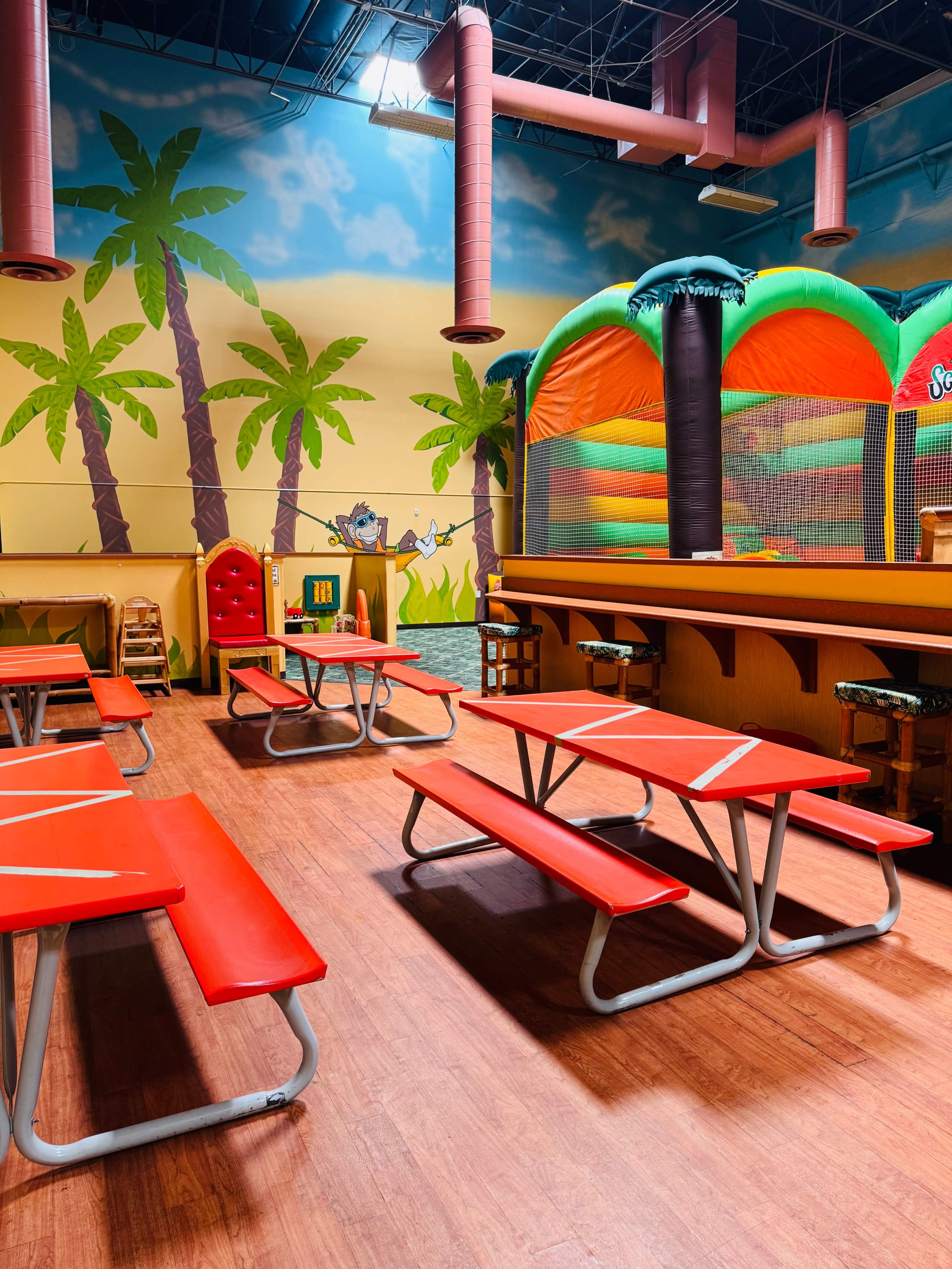 The image shows a colorful indoor play area with green palm tree murals, red picnic-style tables, and a large inflatable bounce house.