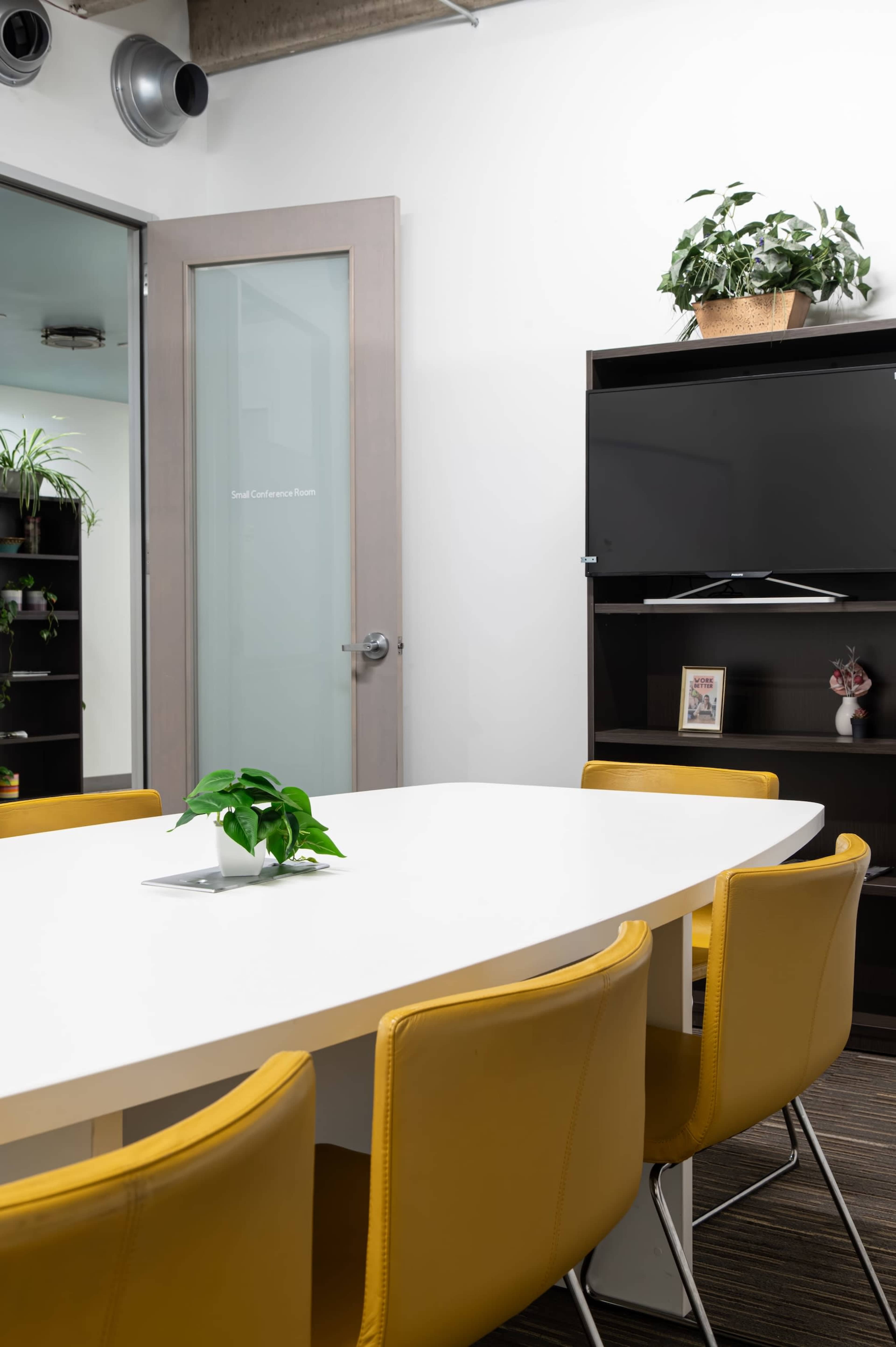 A bright meeting room features a white table surrounded by yellow chairs, with a television mounted on a shelf and plants displayed nearby.