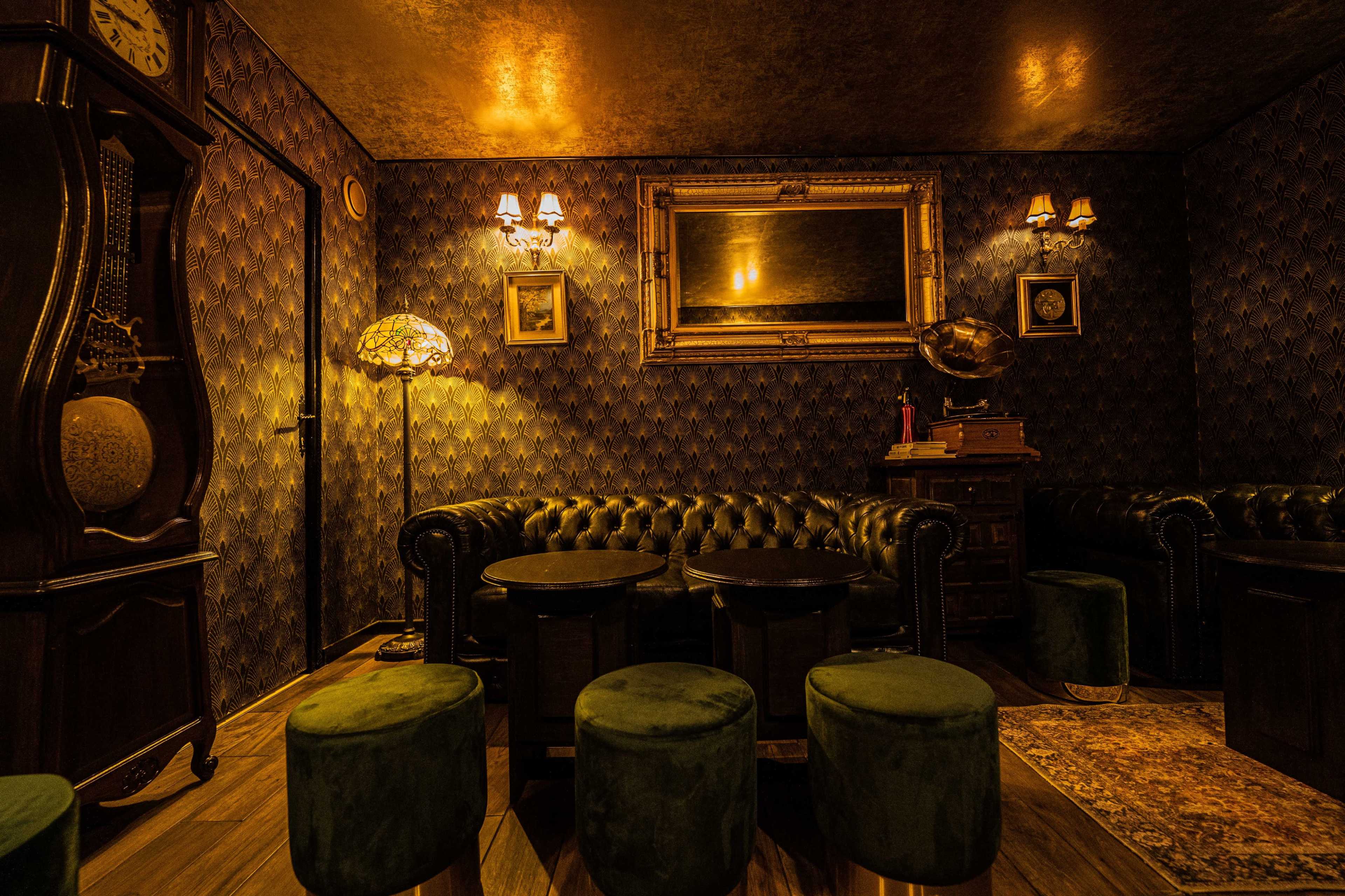 A dimly lit lounge featuring a vintage clock, leather chesterfield sofas, green stools, and decorative wall sconces.