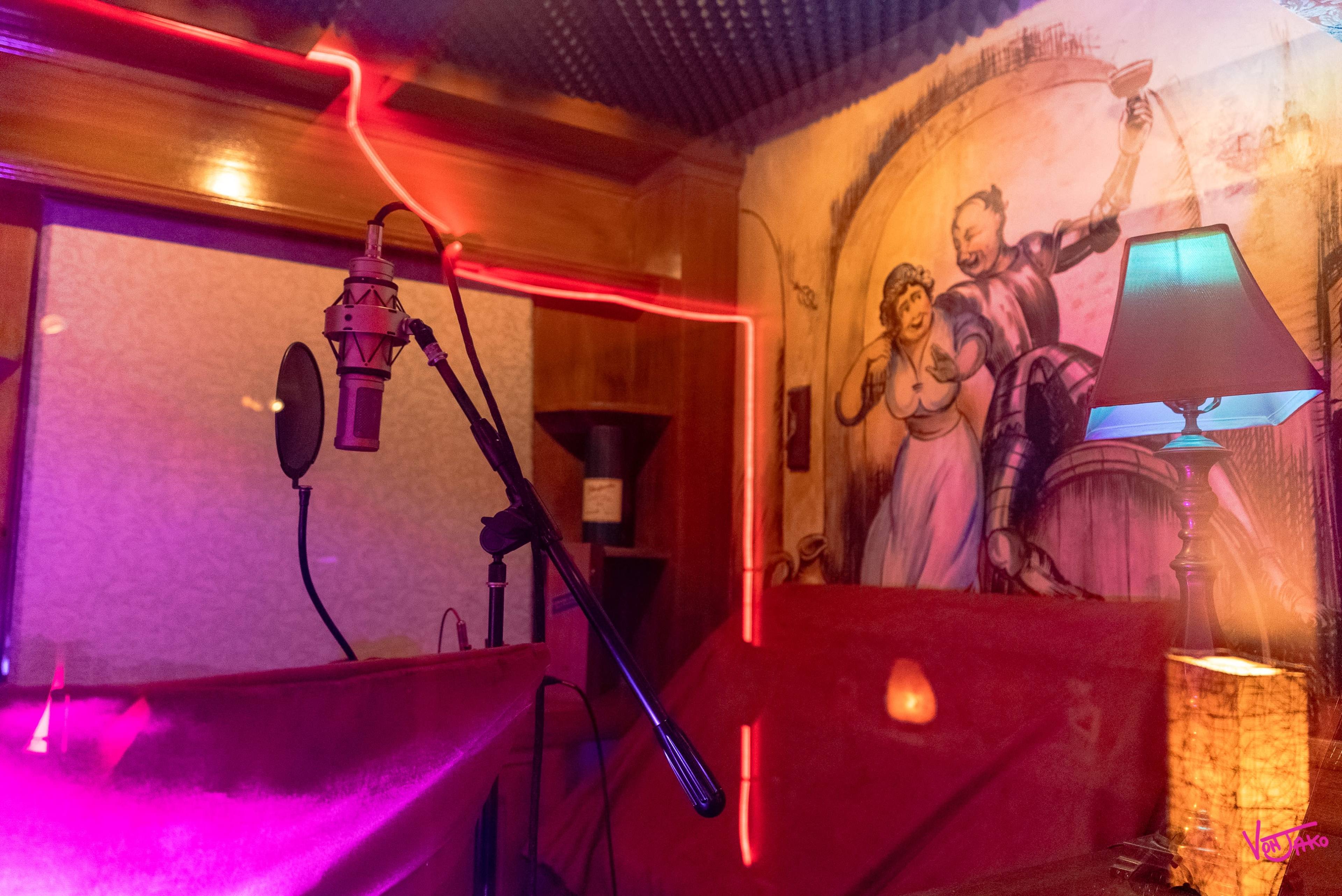 A microphone stands in a recording booth with a colorful lighting design and a mural of a knight and a woman on the wall.