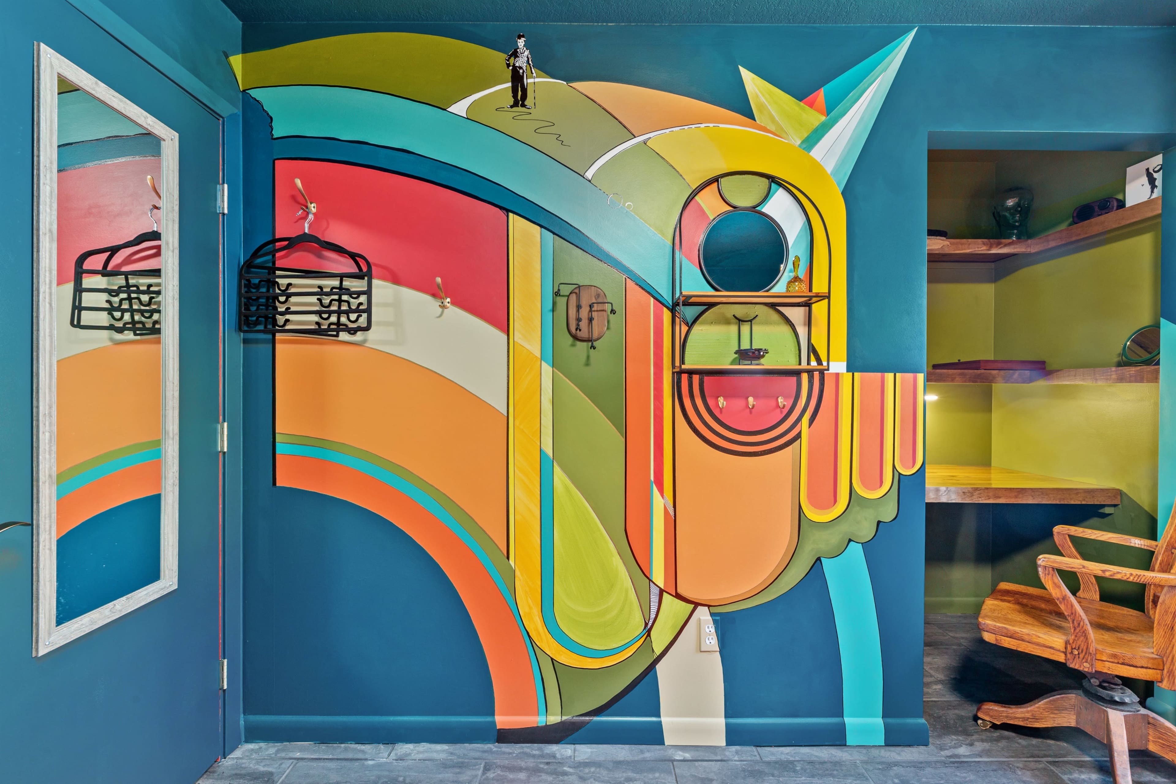 A brightly colored mural on a wall, incorporating abstract shapes and lines in orange, green, and blue, alongside a large mirror and wooden shelf.
