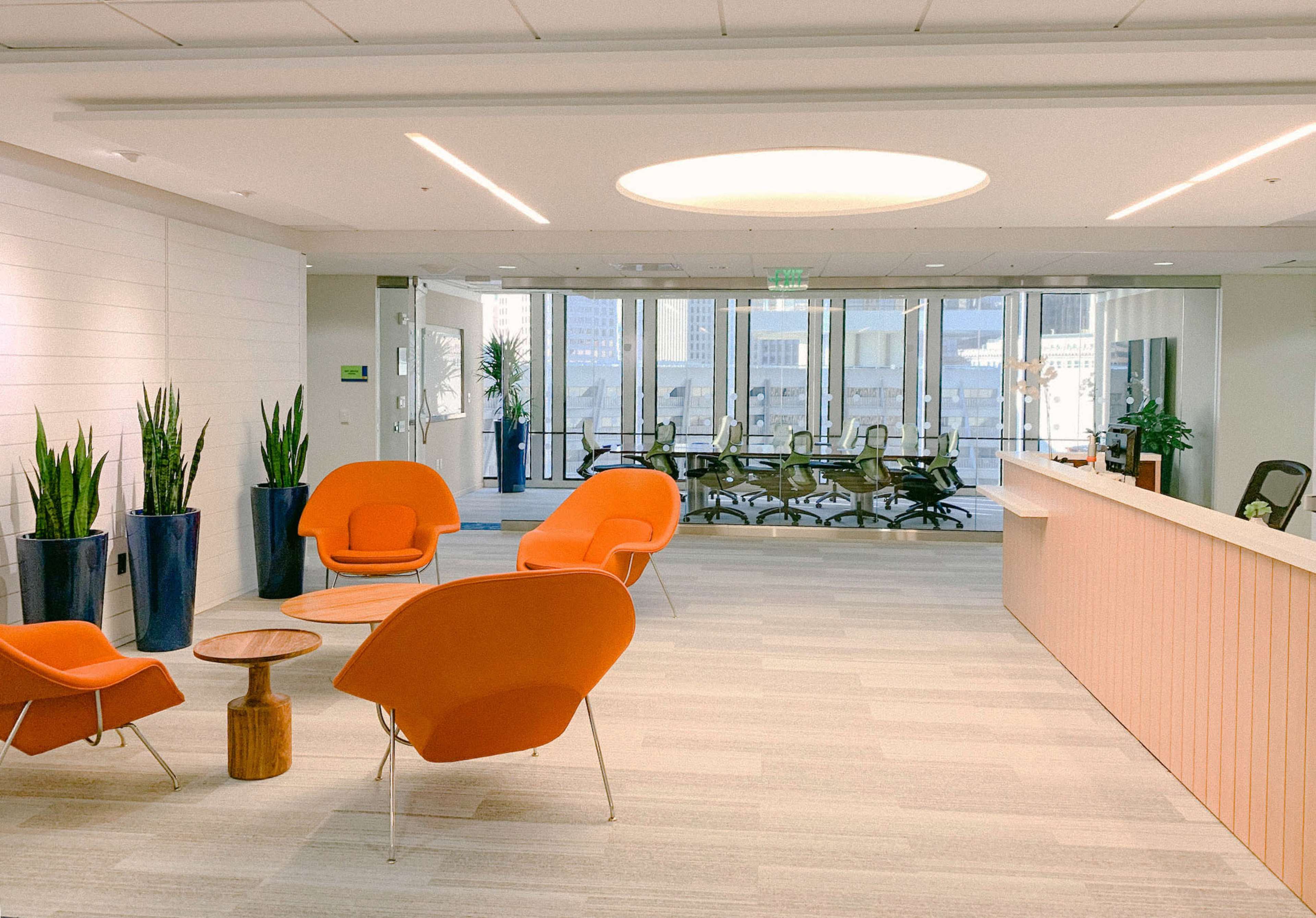 A modern office reception area featuring orange chairs, a wooden table, potted plants, and a glass-walled conference room in the background.