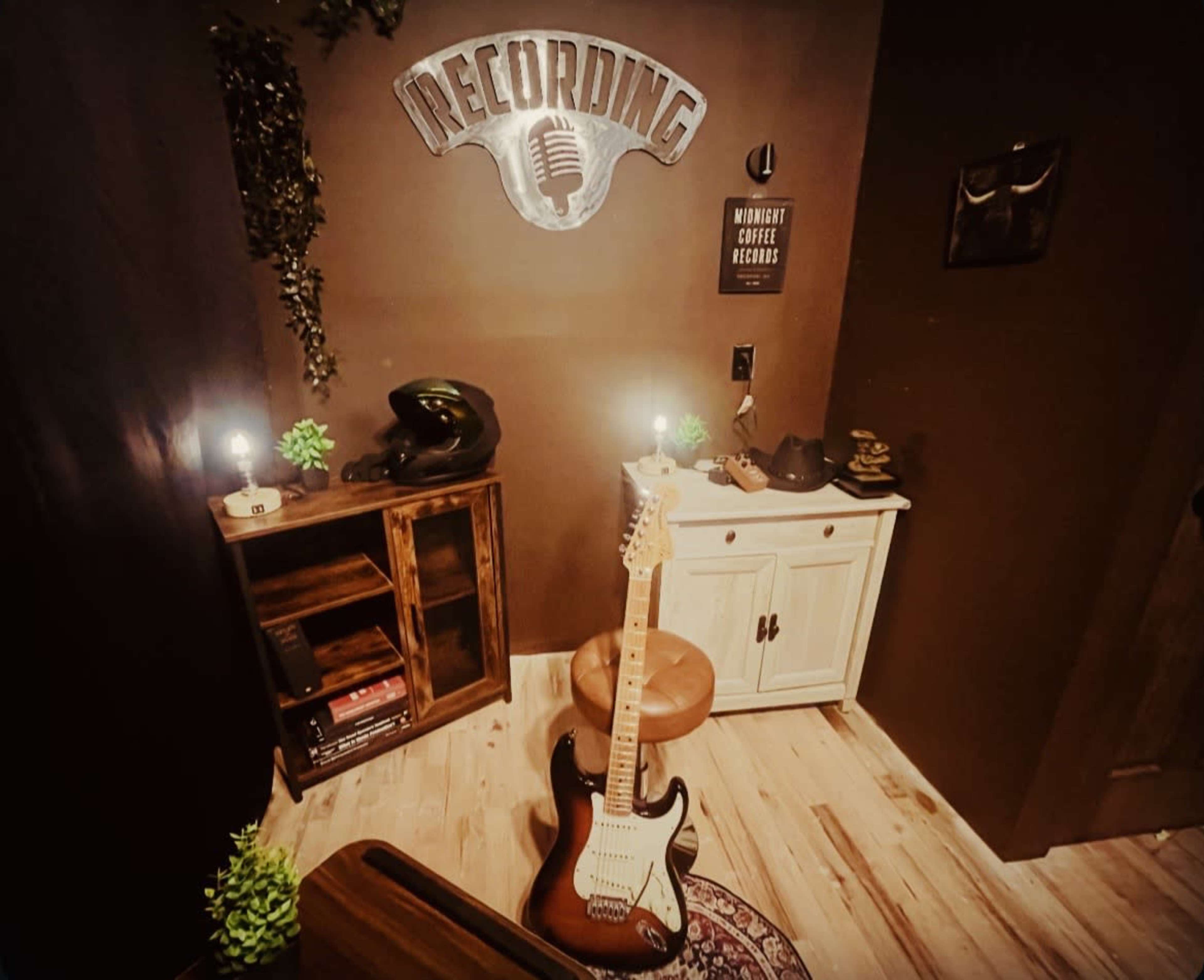 The image shows a small music recording space featuring a guitar, a wooden shelf, and a decorative sign on the wall.