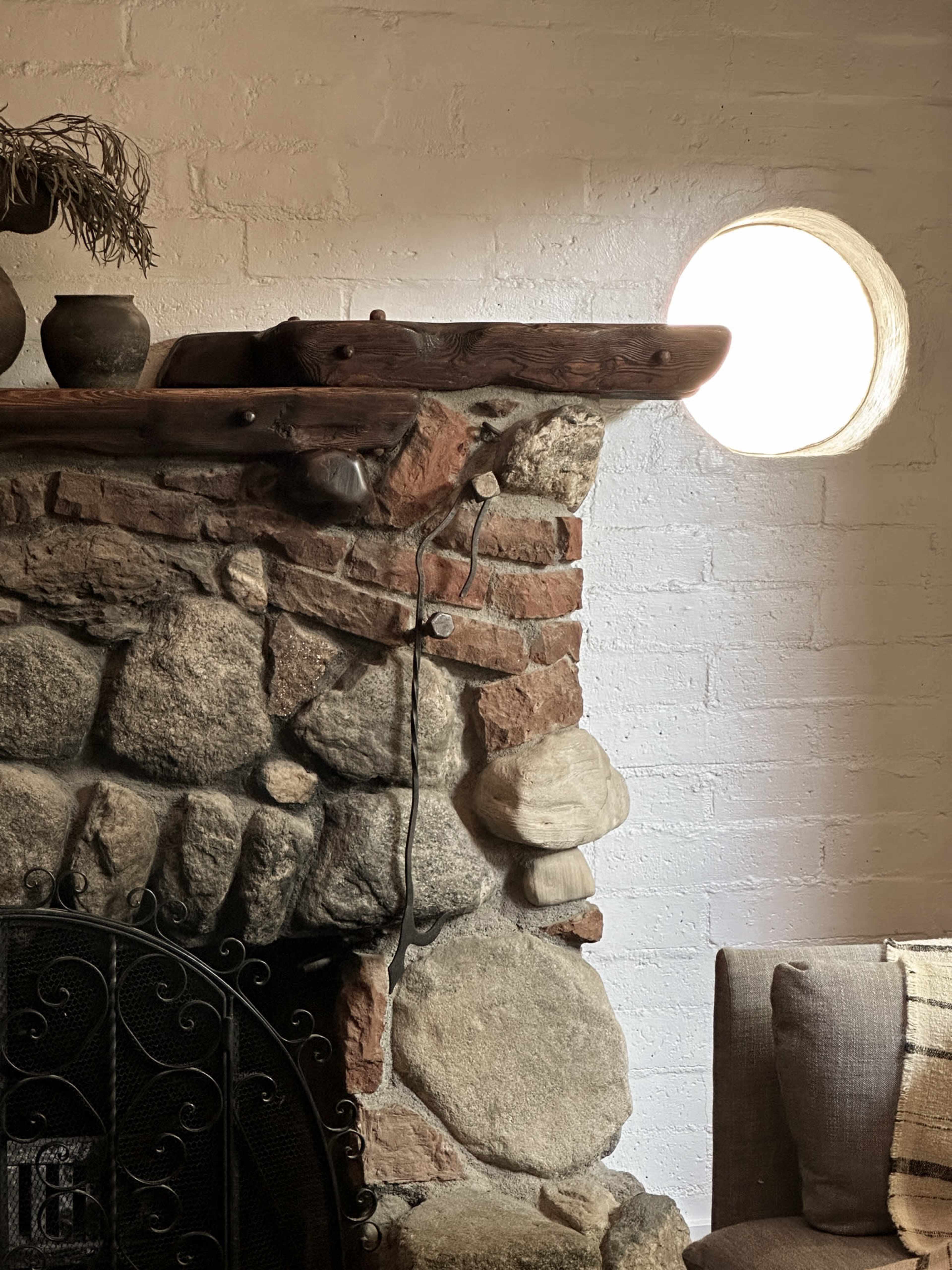 The image shows a rustic stone fireplace with a wooden mantel and a circular window illuminating the space.
