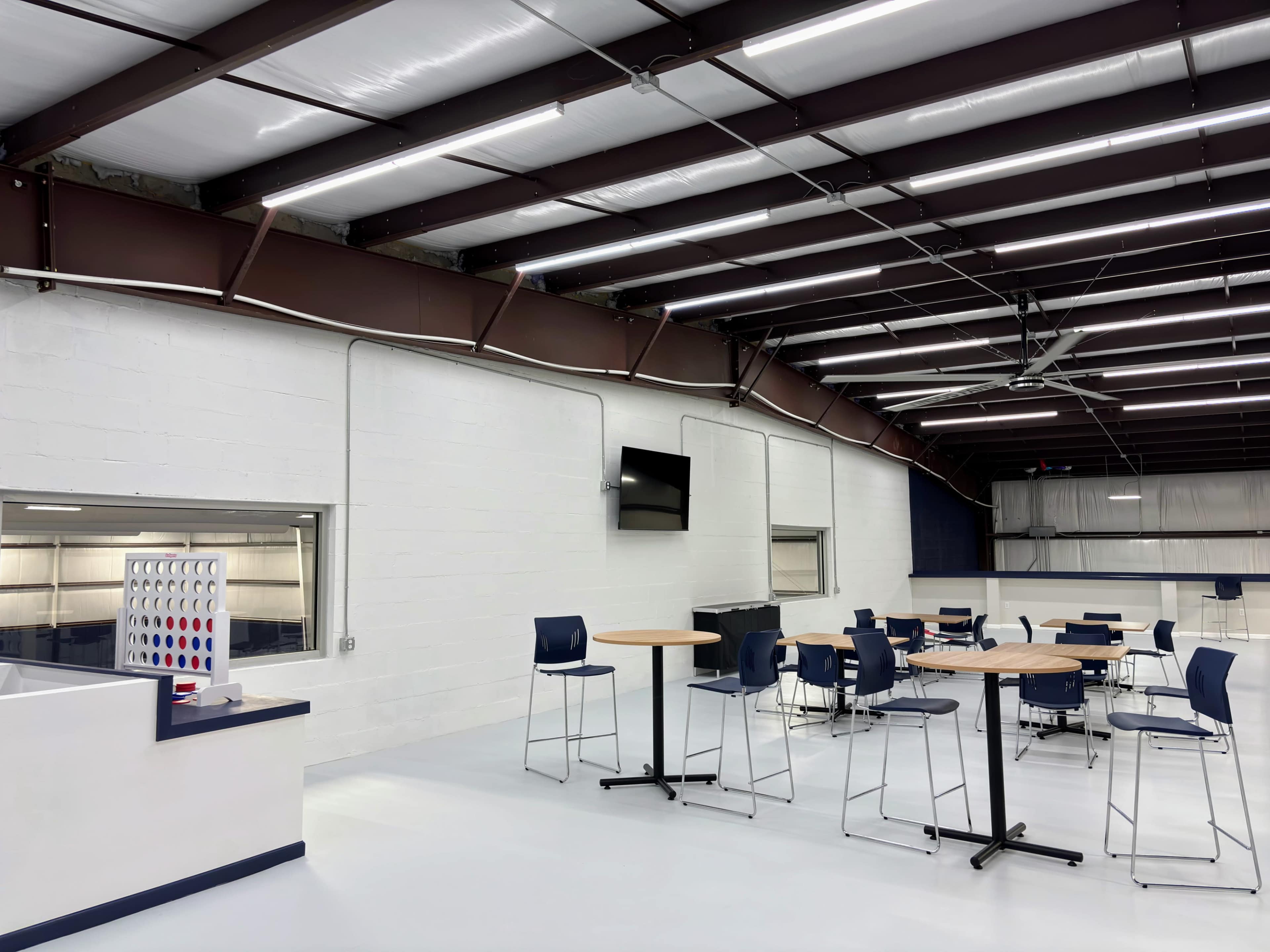 The image shows a modern indoor space with tables and chairs arranged for dining or socializing, equipped with overhead lighting and a large window.