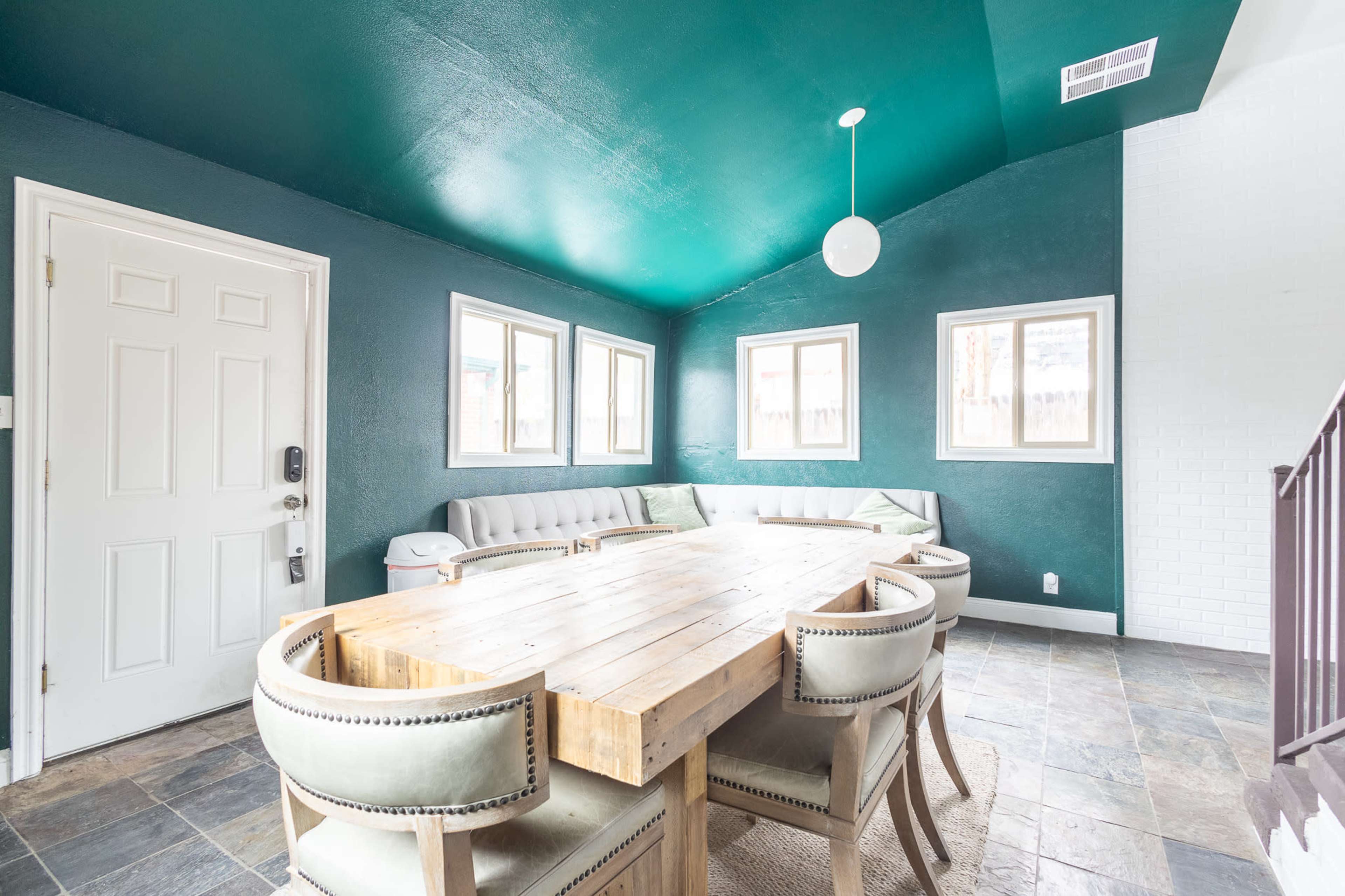 The image shows a modern dining area with a wooden table surrounded by upholstered chairs, featuring a green ceiling and multiple windows letting in natural light.