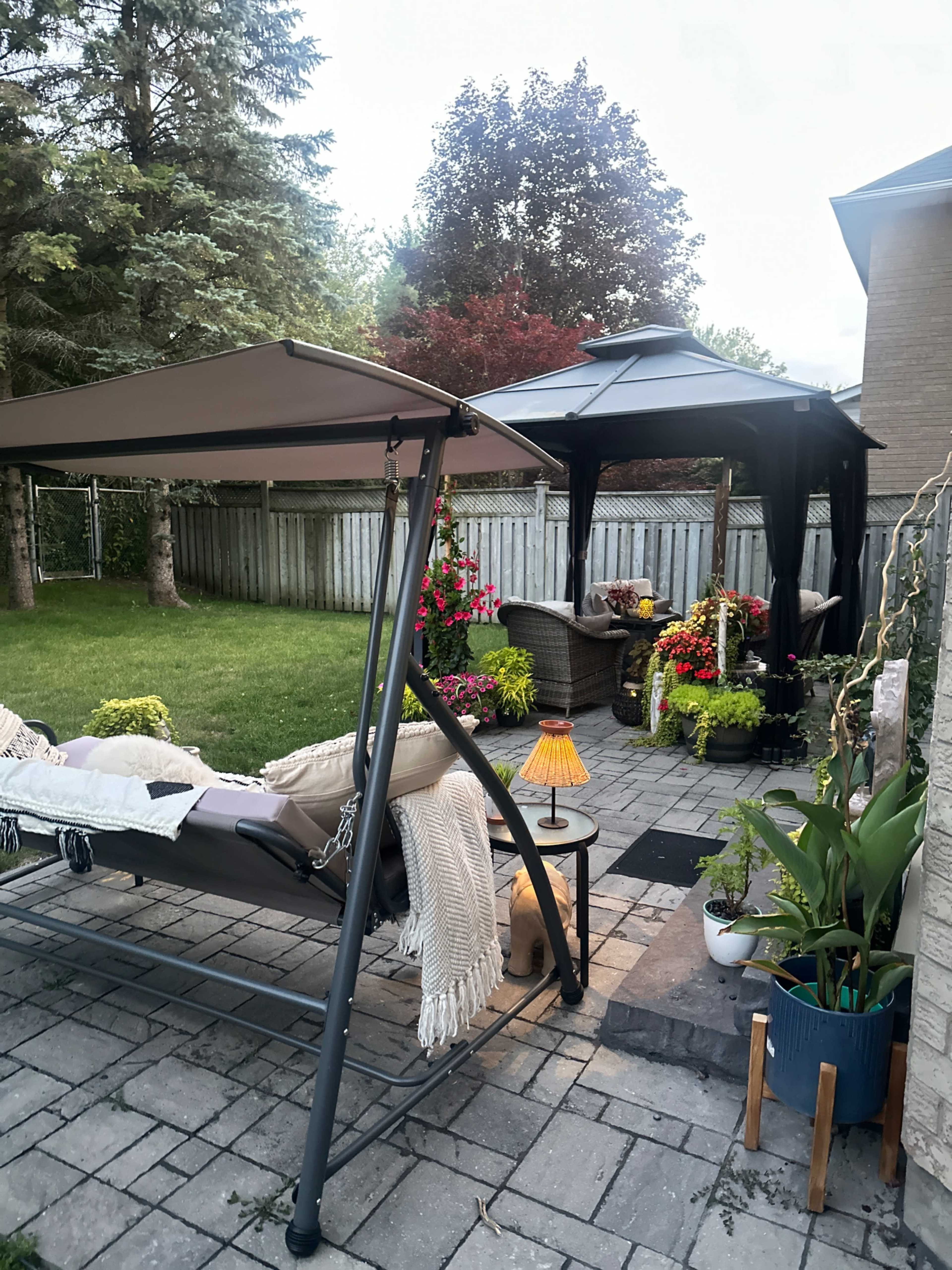 The image shows a backyard scene with a swing chair, a gazebo, potted plants, and colorful flower arrangements along a stone patio.