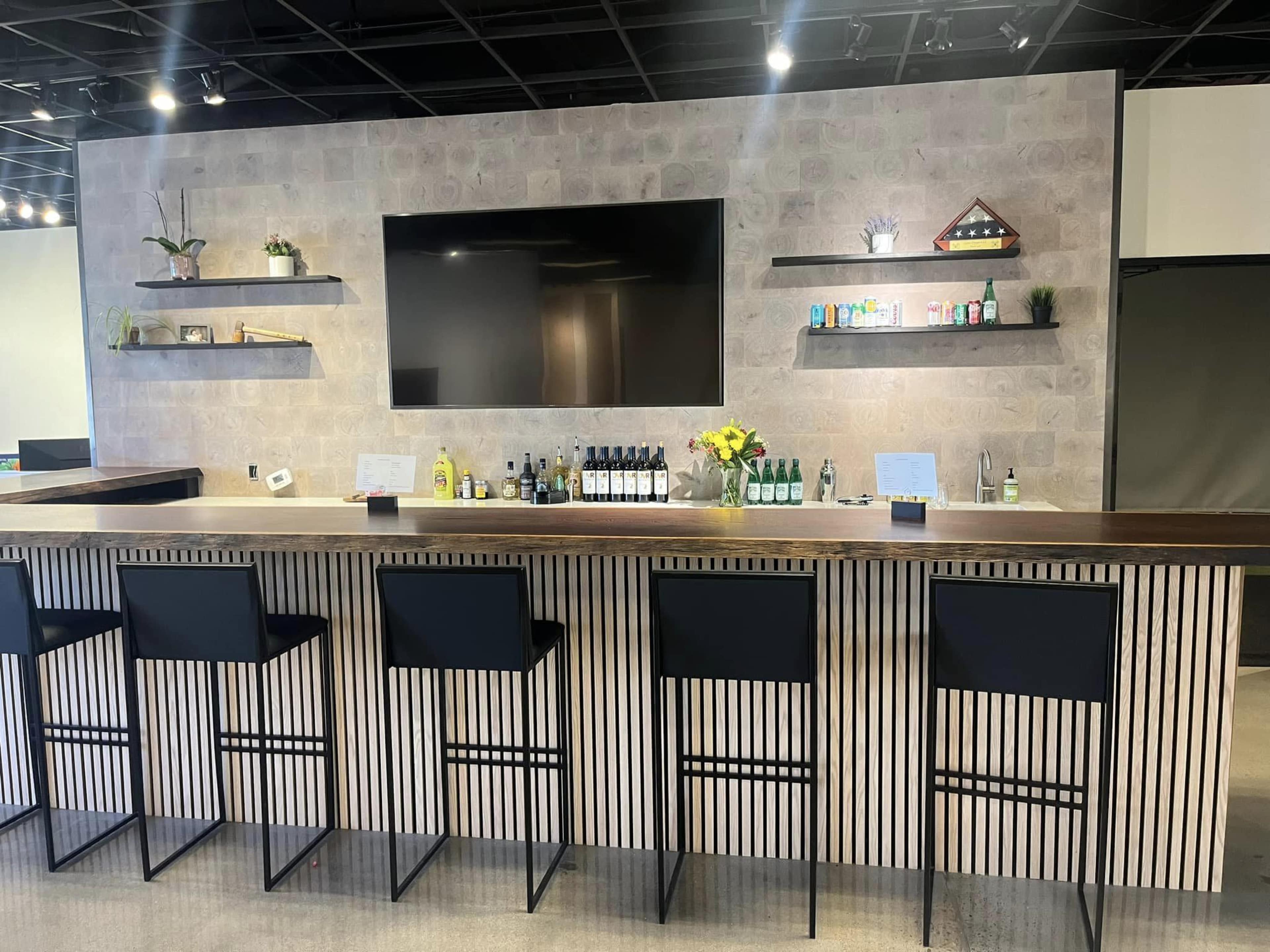 The image shows a modern bar with a long, wooden counter, black high chairs, a mounted television, and shelves displaying various bottles and decorative items against a textured wall.