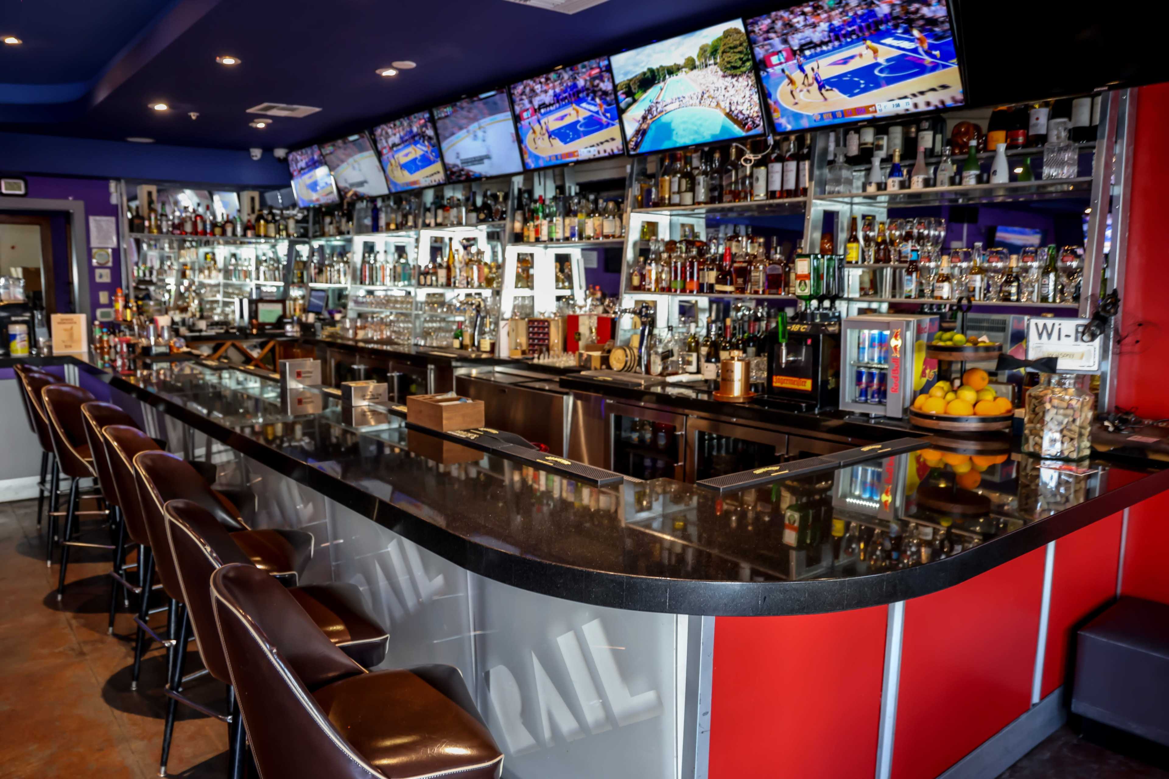 The image shows a lively bar interior with a long black countertop, multiple televisions displaying sports, and a well-stocked back shelf of liquor bottles.