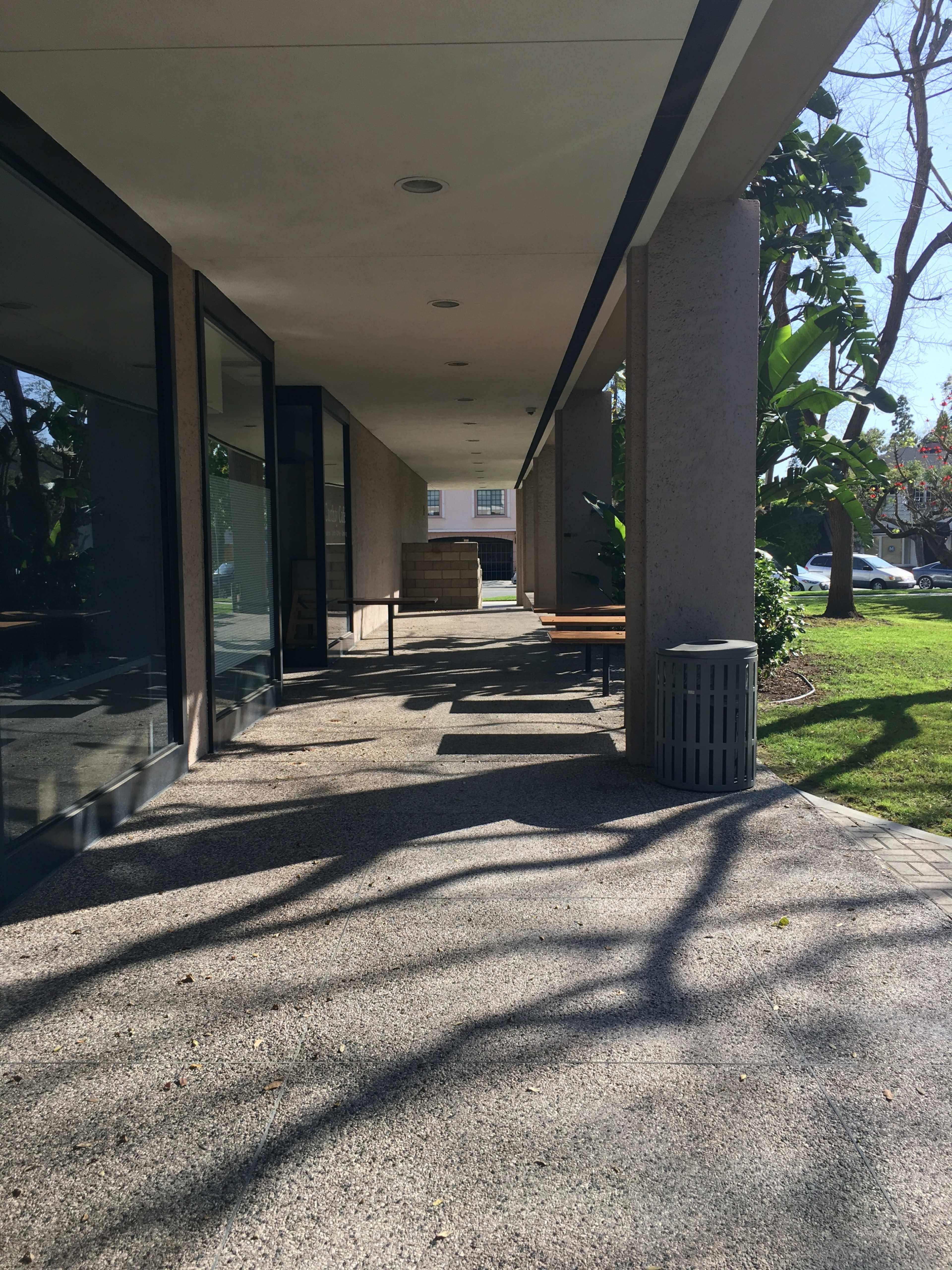 The image shows a walkway beside a building with benches and a trash can, surrounded by trees and grass.