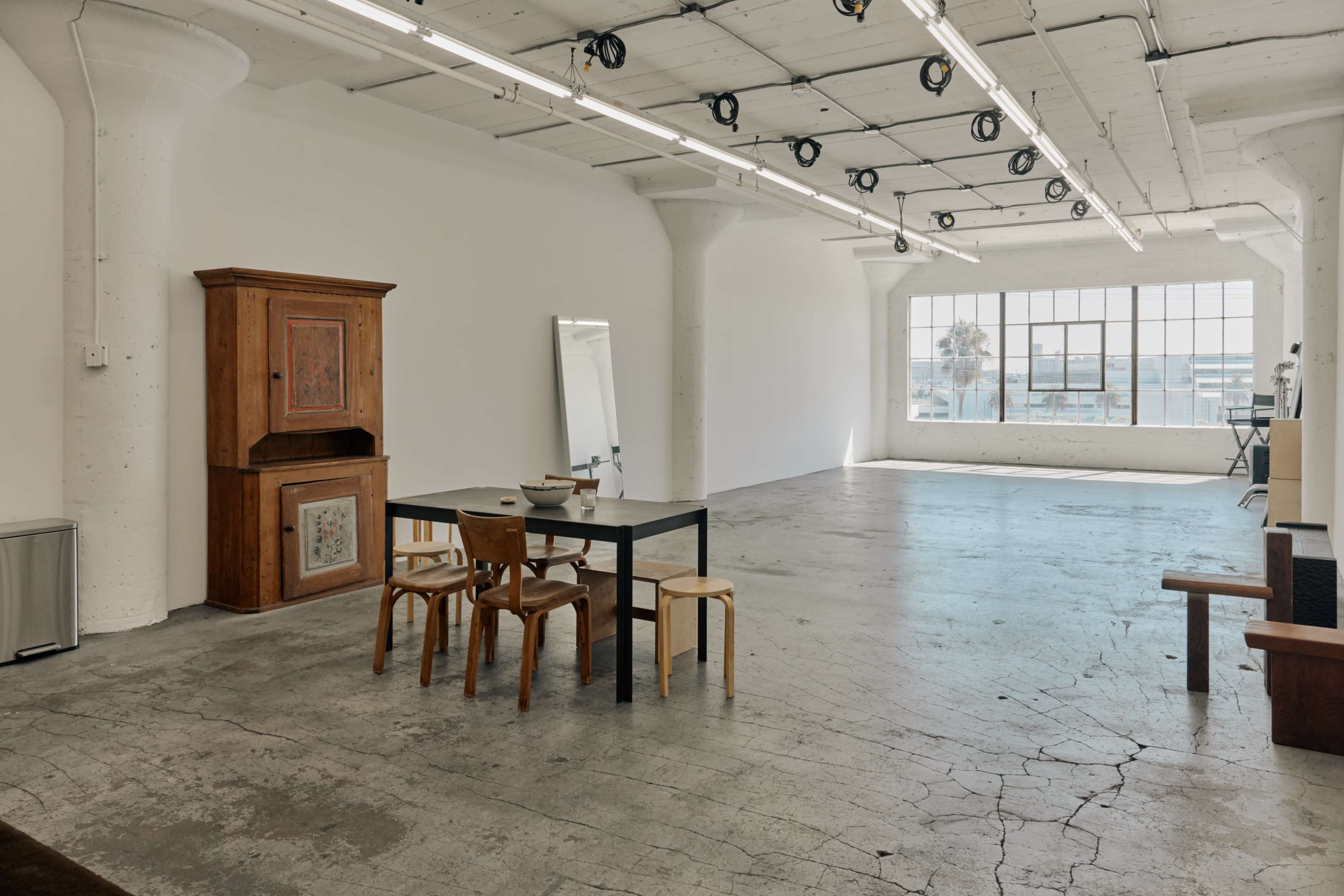 The image shows a spacious, empty room with concrete flooring, featuring a wooden cabinet, a table with chairs, and large windows letting in natural light.
