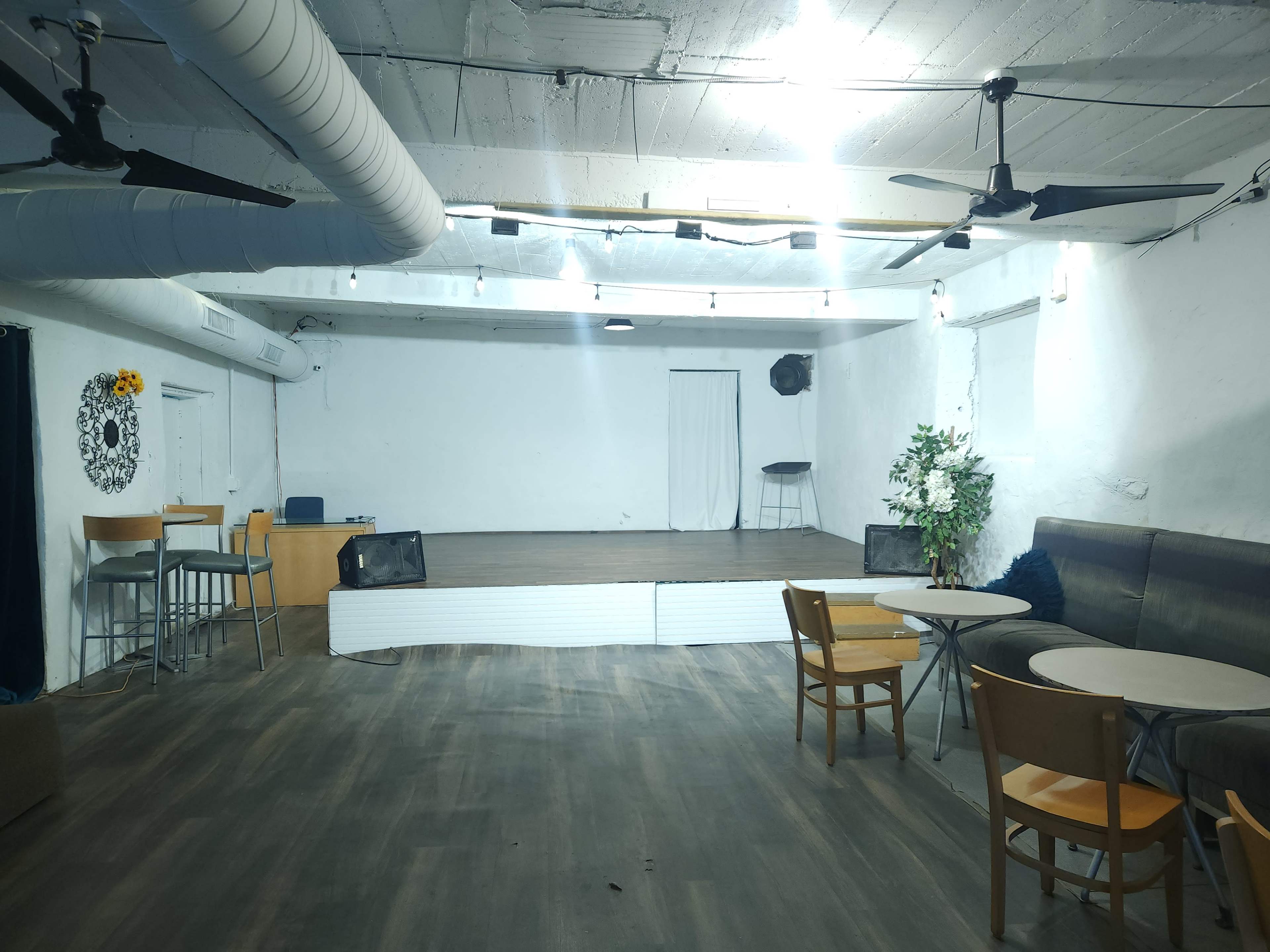 An empty event space with a wooden stage, scattered tables, chairs, and a few decorative elements on the walls.