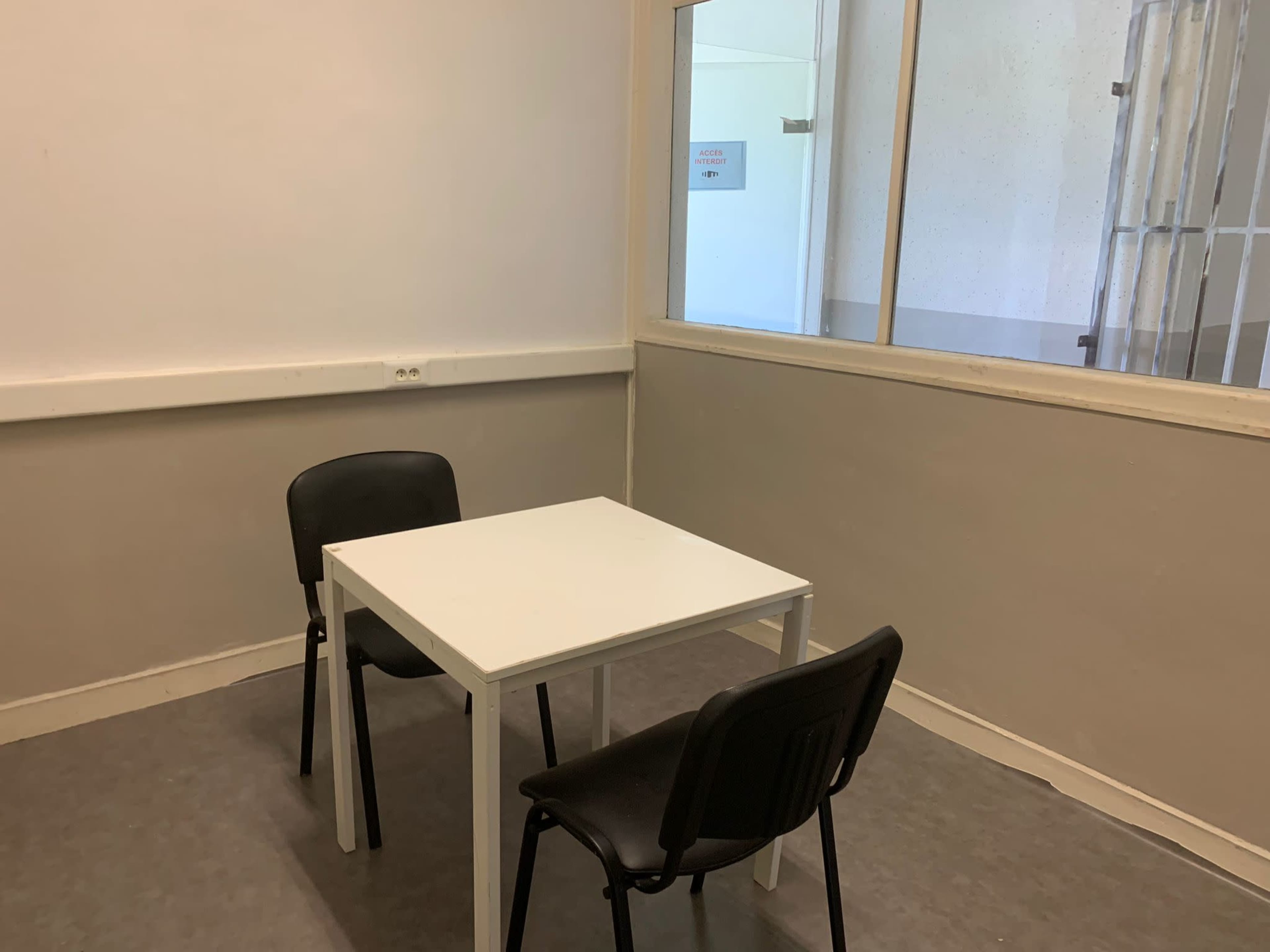 A small room with a white table and two black chairs, featuring a window with a barred view.