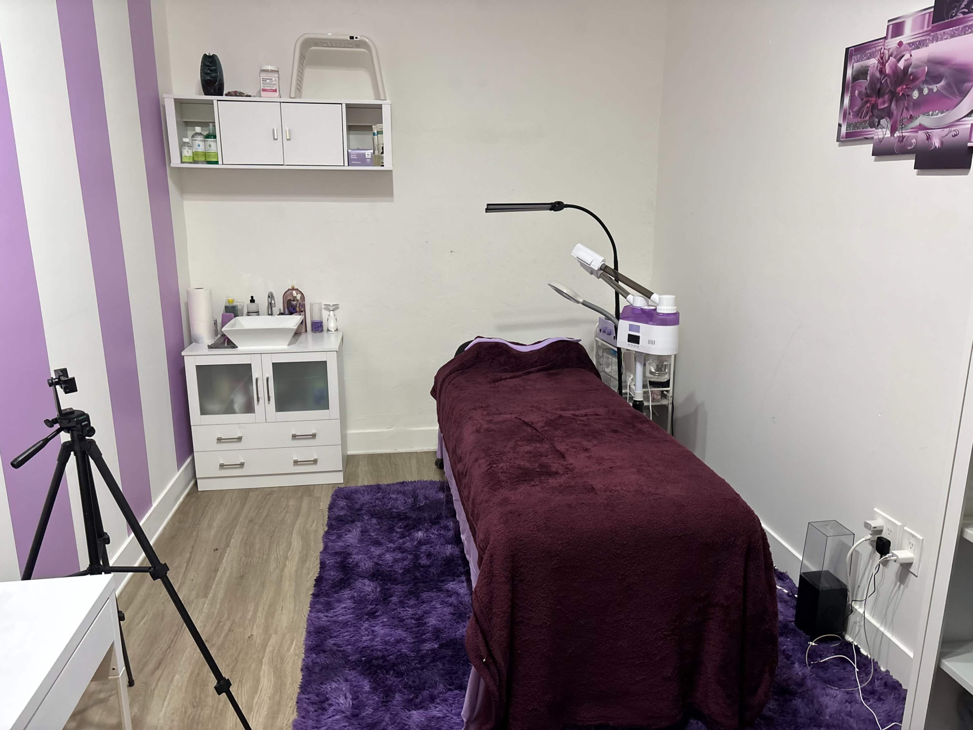 The image shows a treatment room with a purple-themed decor, featuring a treatment bed, storage cabinets, and a camera tripod set up in the corner.