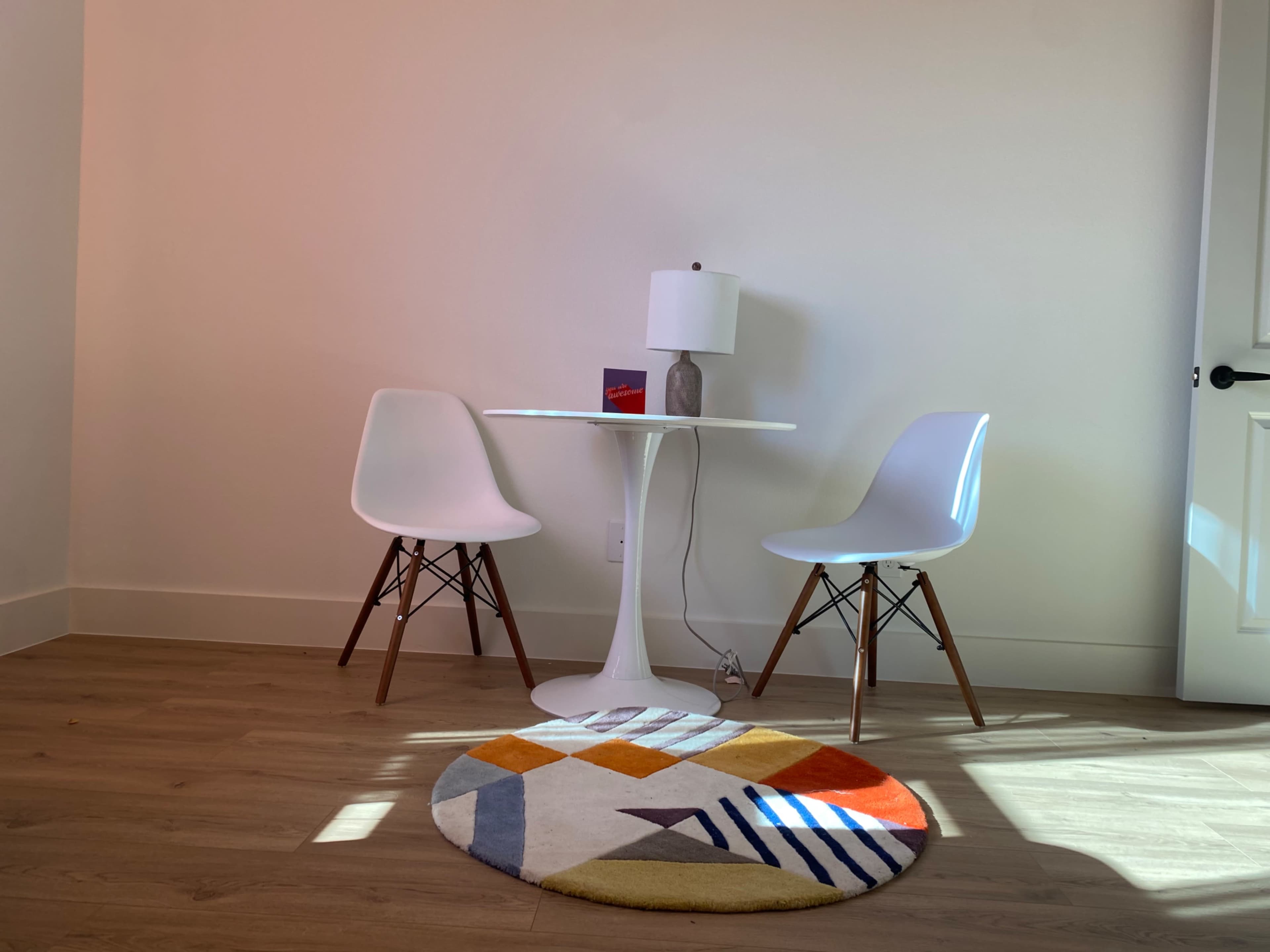 A small table with two white chairs and a lamp stands on a patterned rug in a minimalistic room with light-colored walls.