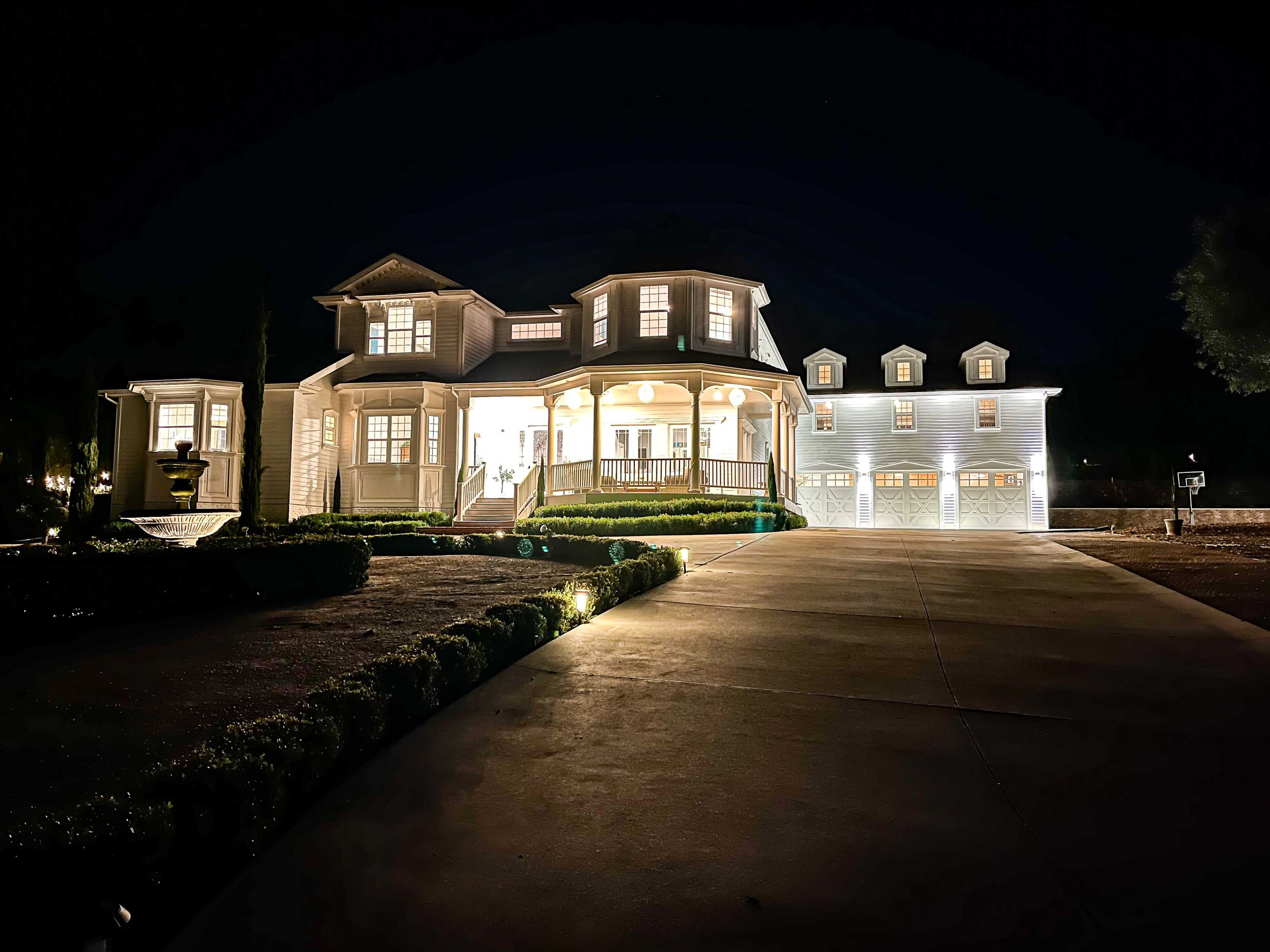 A large, well-lit house with multiple stories and a two-car garage stands prominently against the night sky, surrounded by neatly manicured landscaping.