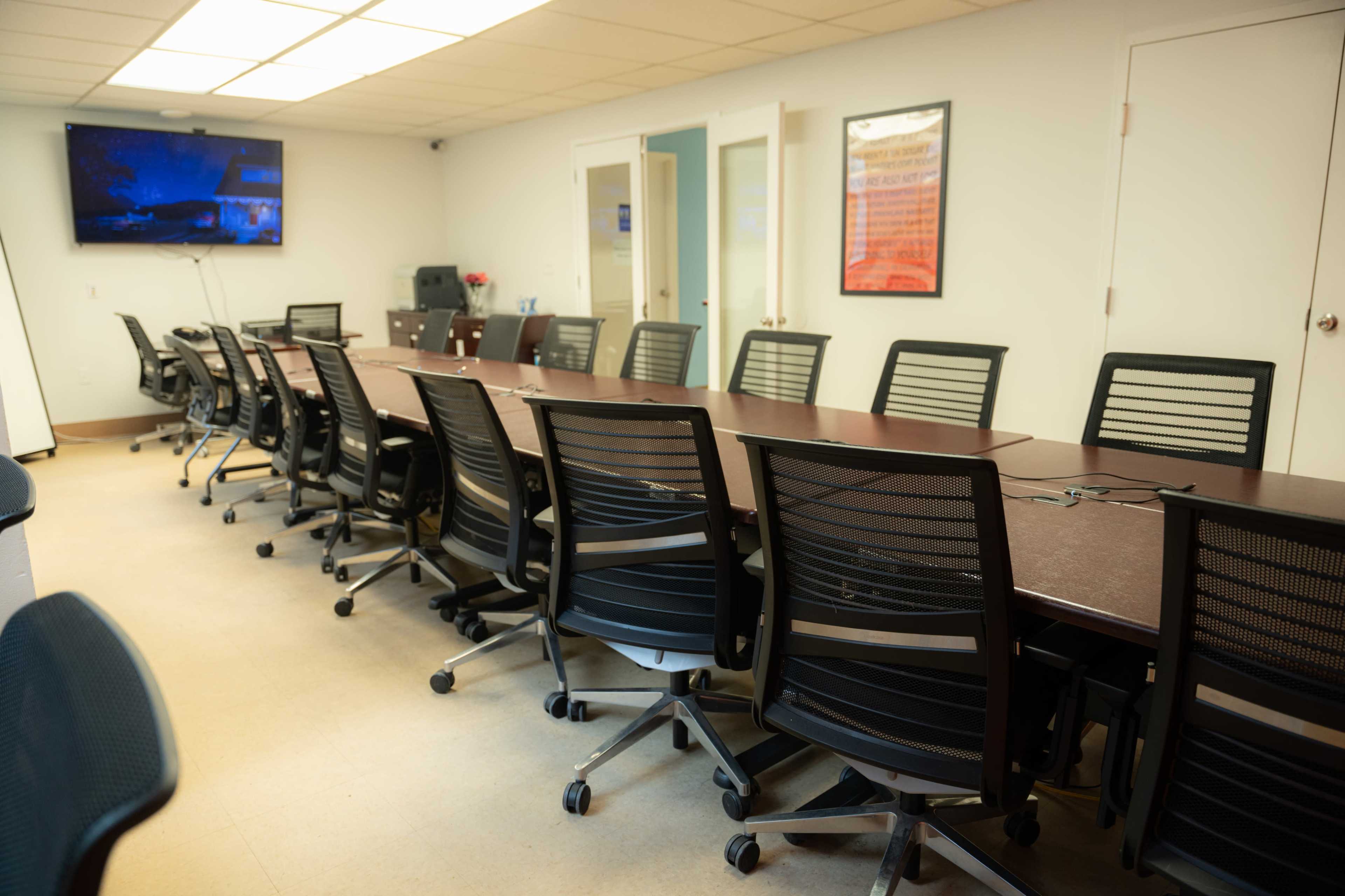 A conference room with a long table surrounded by numerous black office chairs and a wall-mounted television.