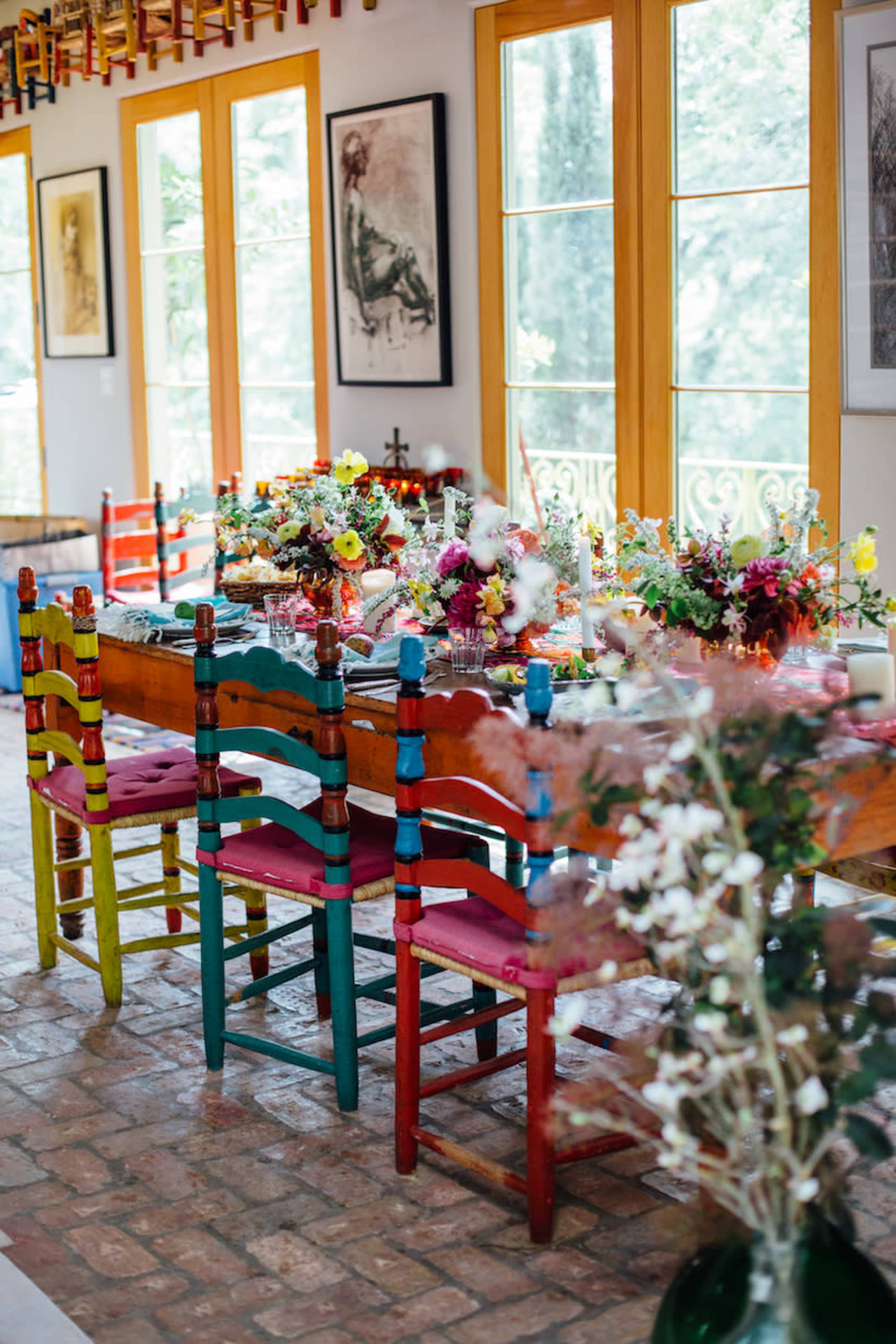 A long wooden dining table is set in a brightly lit room, surrounded by colorful chairs and adorned with floral arrangements.