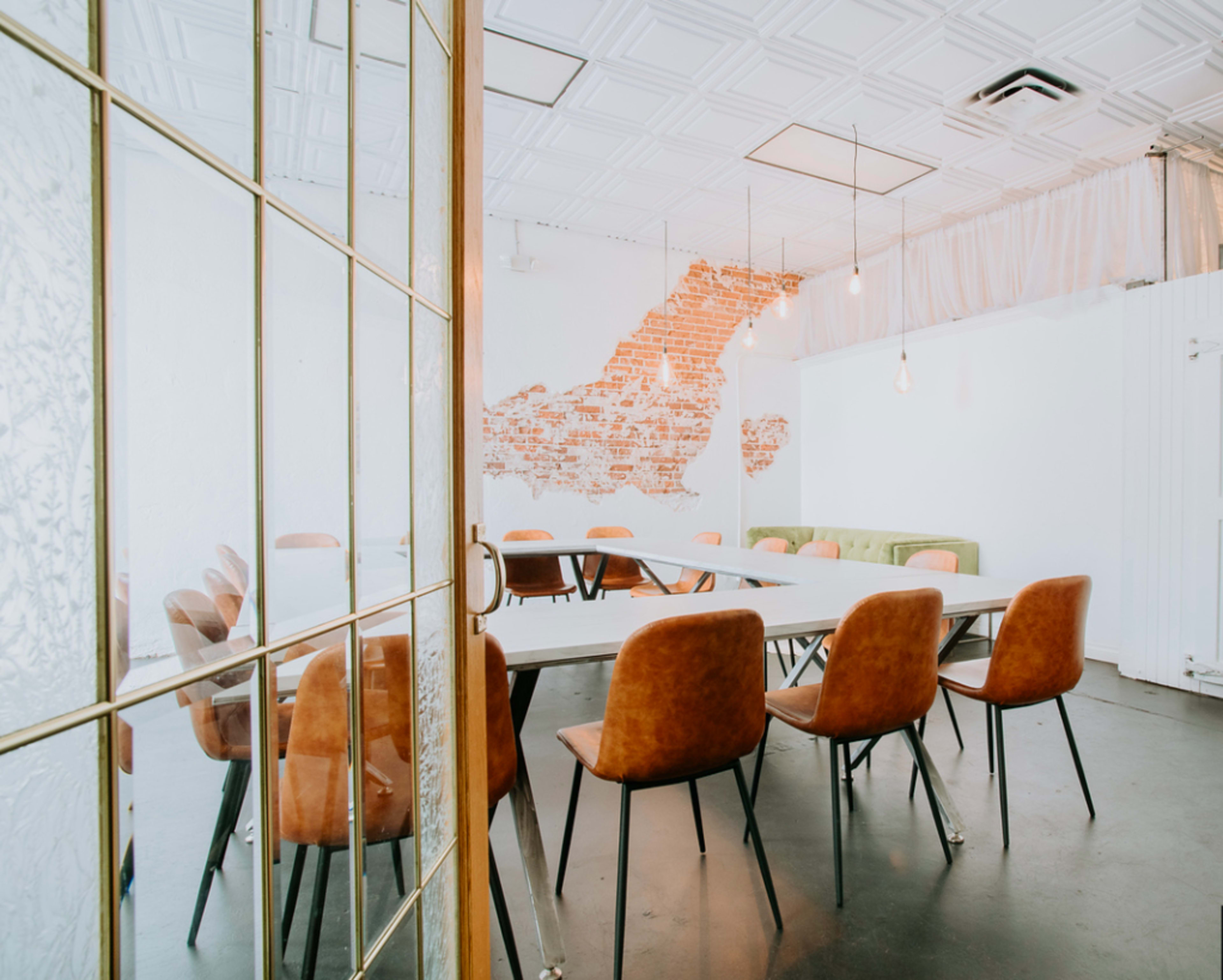 A modern meeting room features a long table surrounded by brown chairs, with exposed brick on one wall and pendant lights hanging from the ceiling.