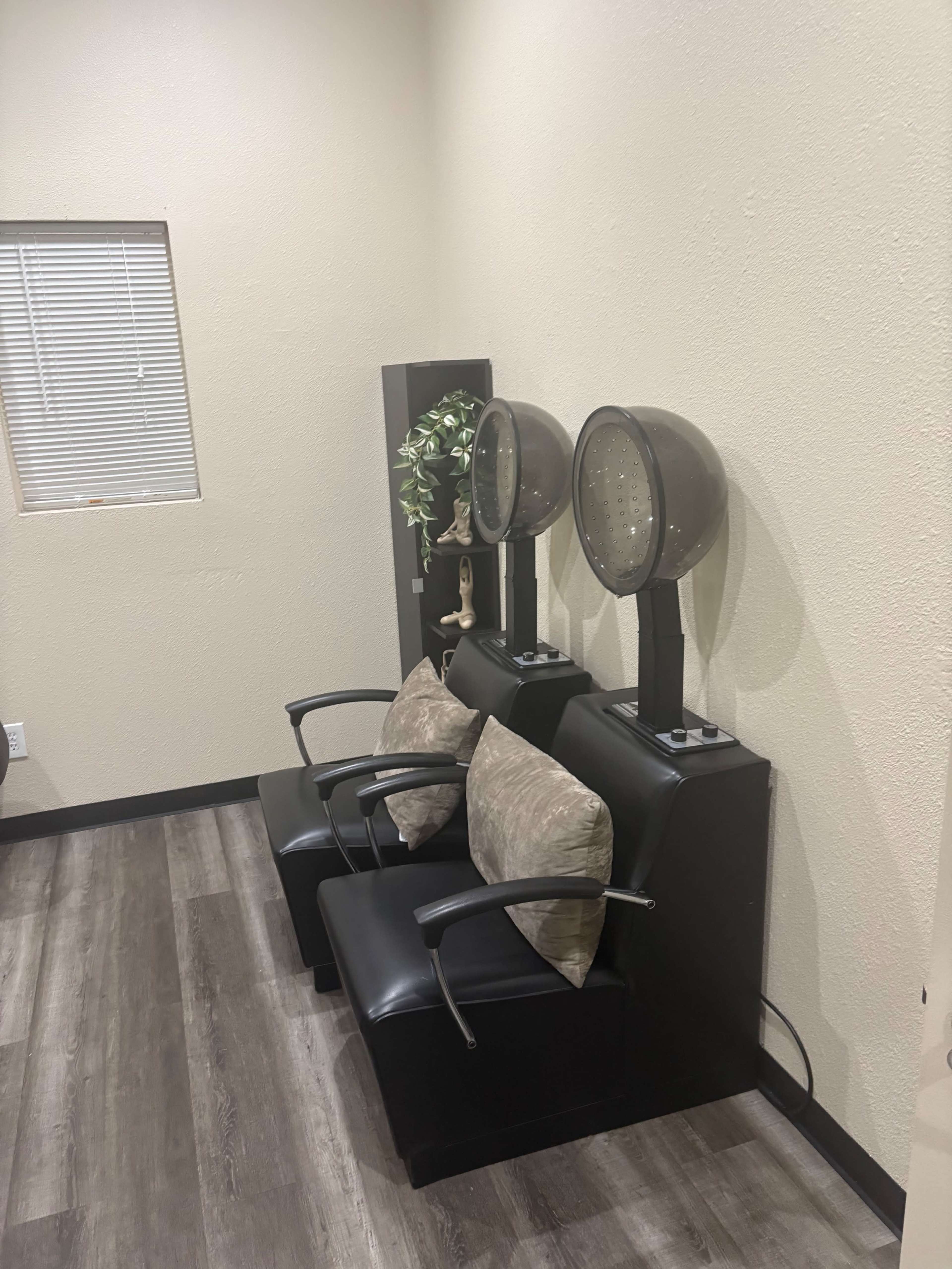 The image shows two black salon chairs with decorative pillows positioned beside two hooded hair dryers against a plain wall.