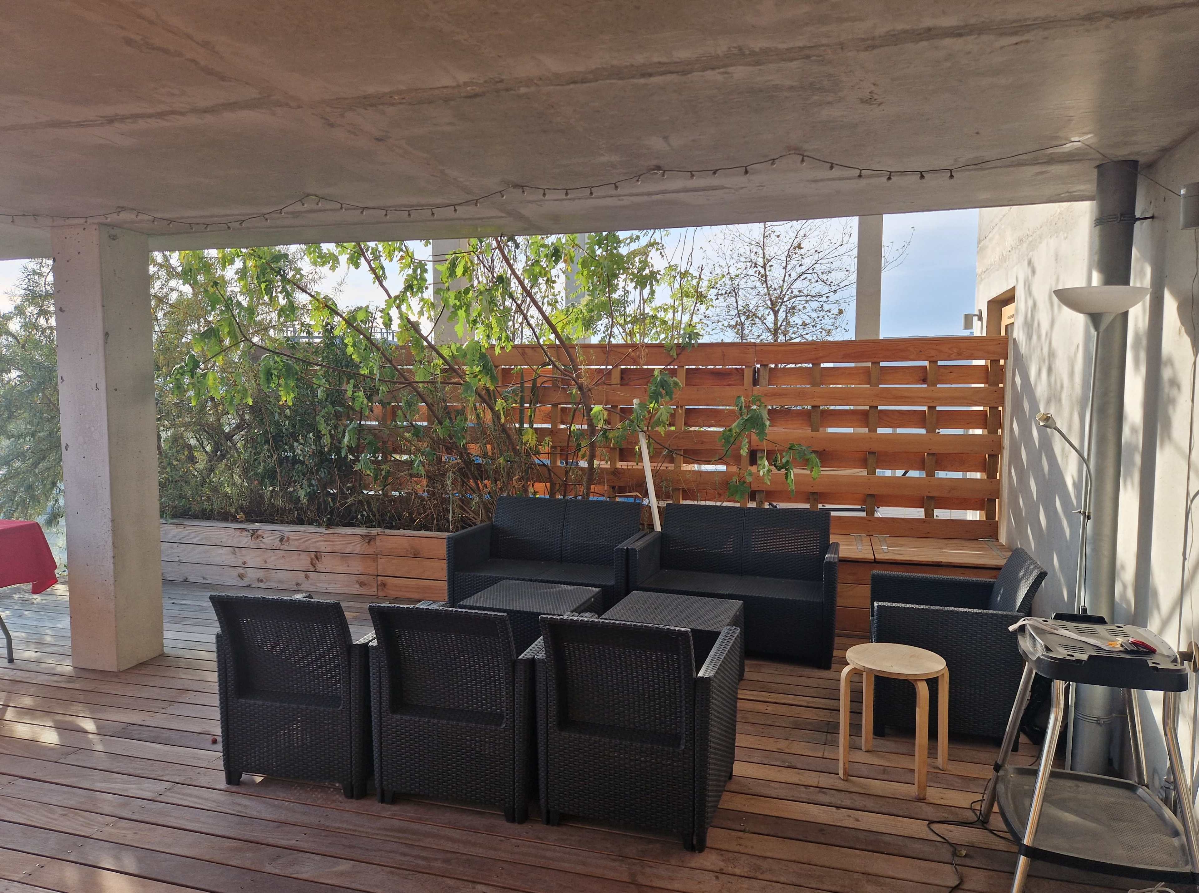 The image shows a covered outdoor patio area furnished with a black wicker seating set and a small wooden table, surrounded by planters and a wooden privacy screen.