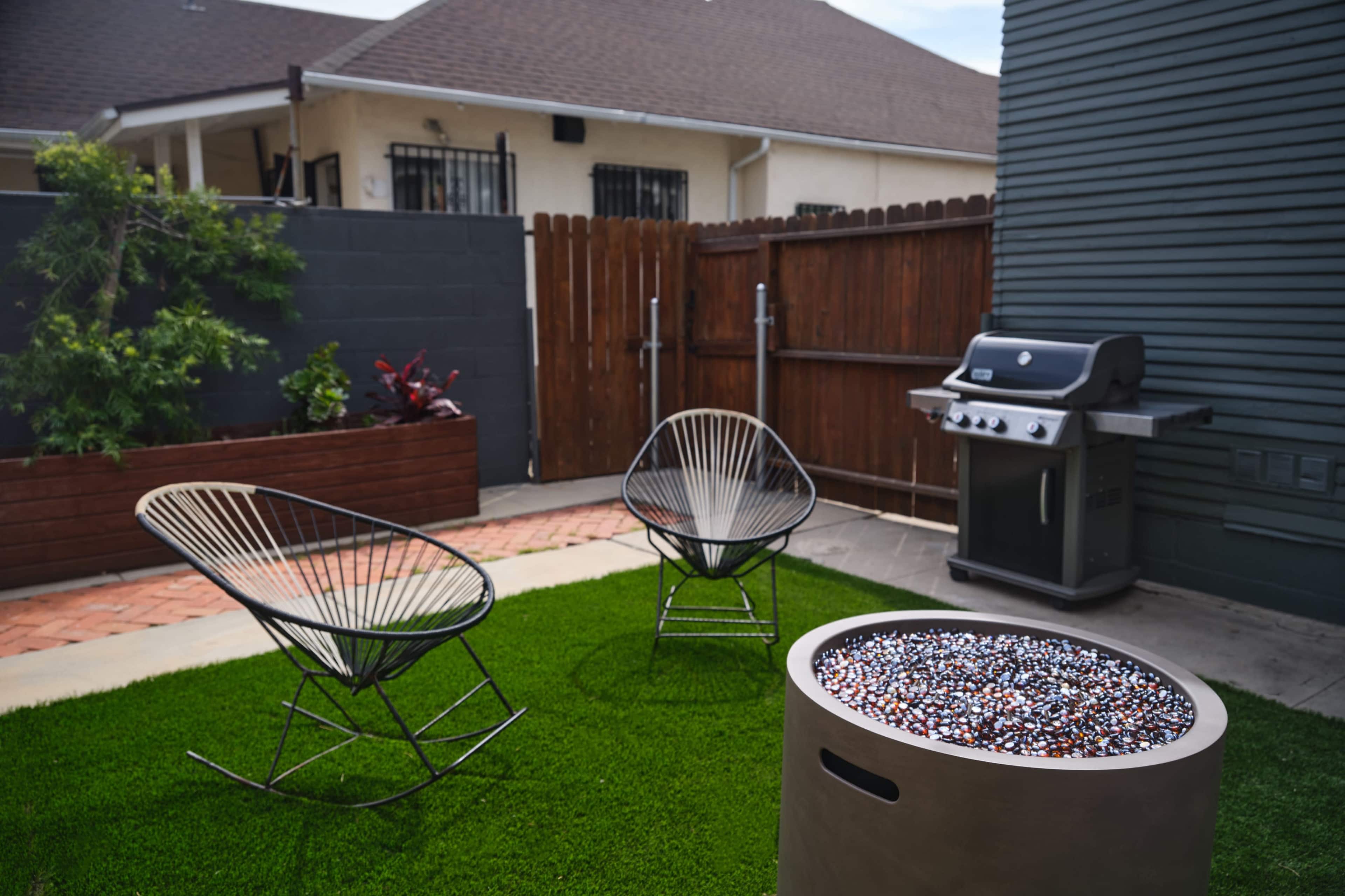The image shows a small outdoor area featuring two wire chairs, a fire pit filled with decorative stones, a barbecue grill, and a wooden fence in the background.