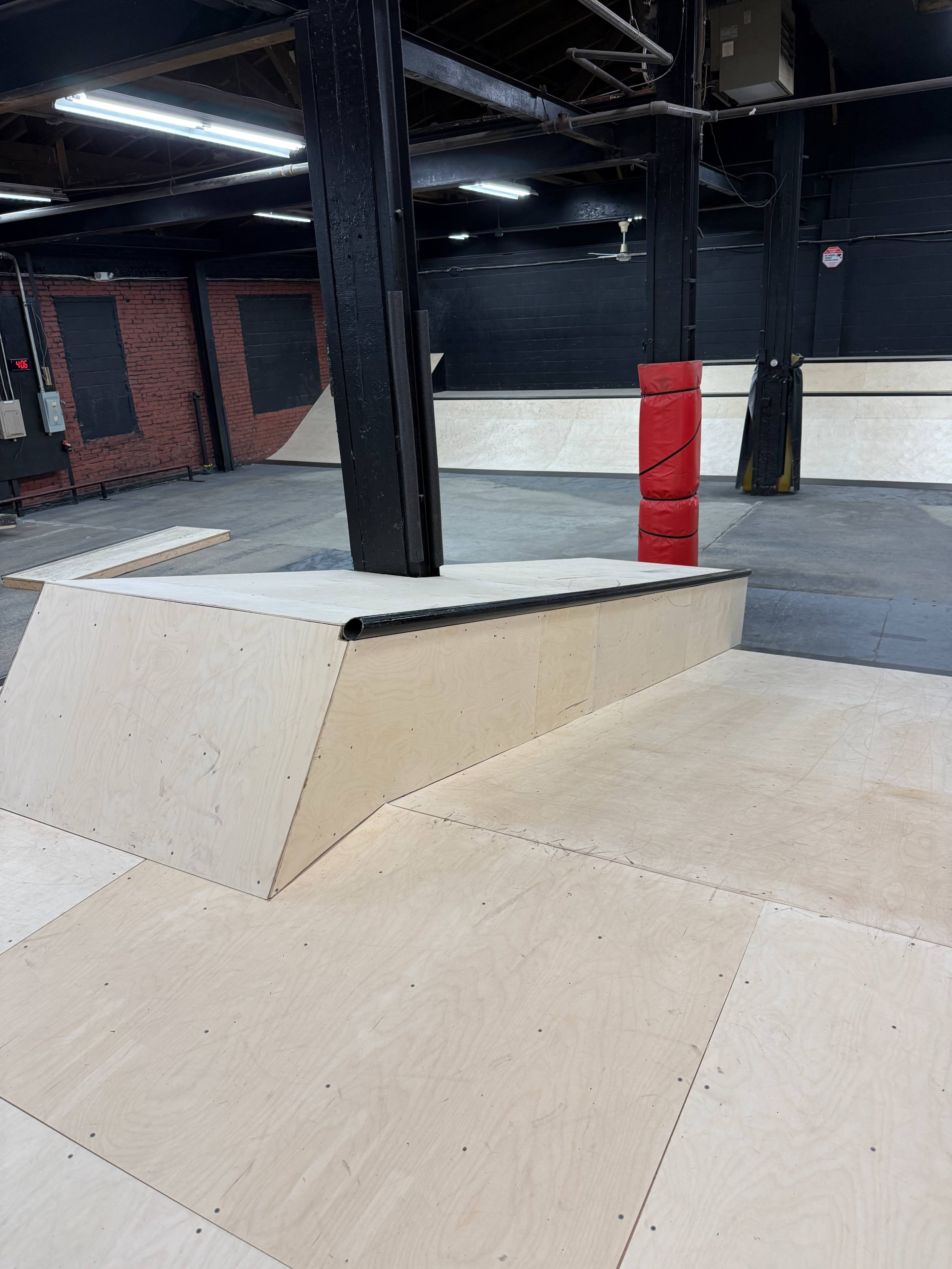 The image shows a skatepark interior featuring wooden ramps and a flat surface, with a red safety post in the foreground.