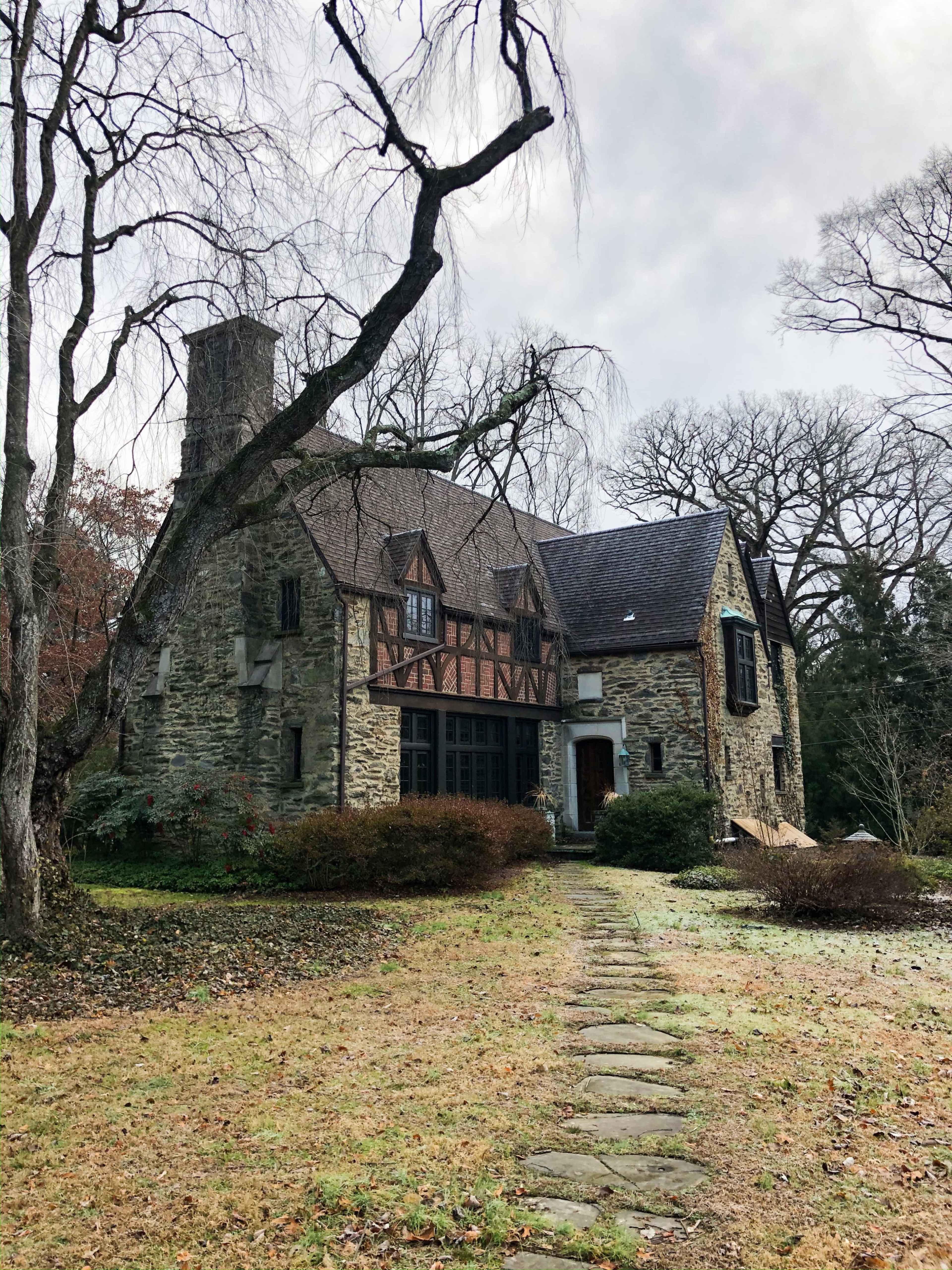 A stone house with a steeply pitched roof and a walkway lined with stones is surrounded by a grassy area and bare trees.